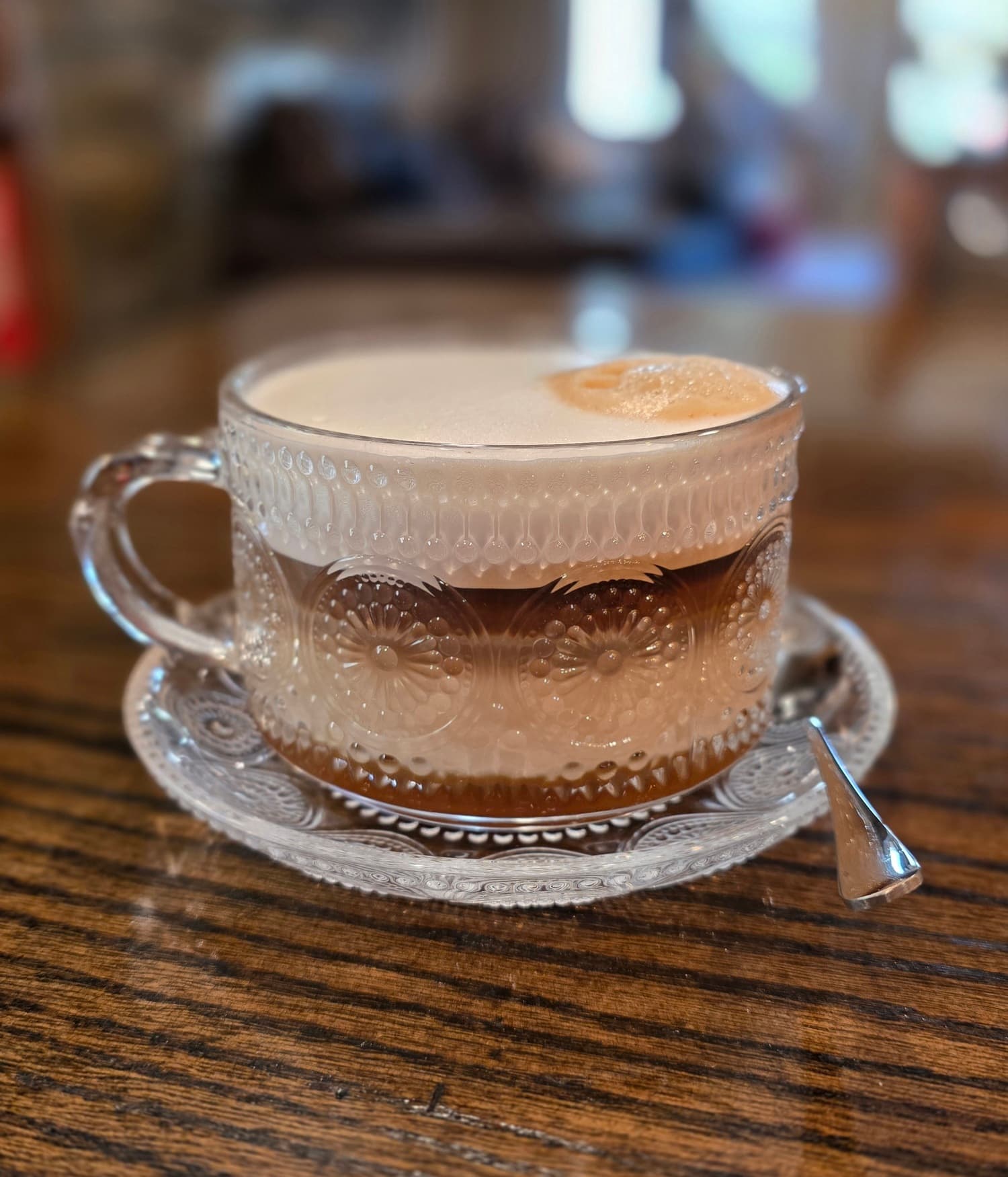 A decorative glass cup filled with layered coffee and topped with foam, resting on an ornate saucer.