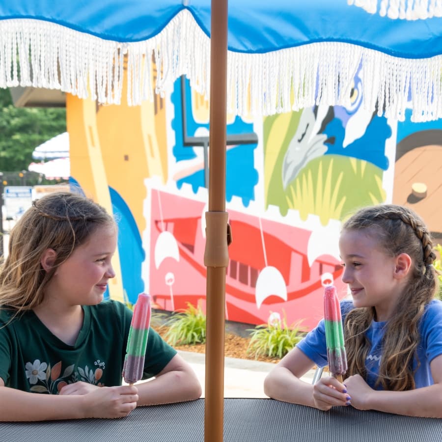 Two girls sit under an umbrella, enjoying colorful popsicles while smiling at each other in front of a vibrant mural.