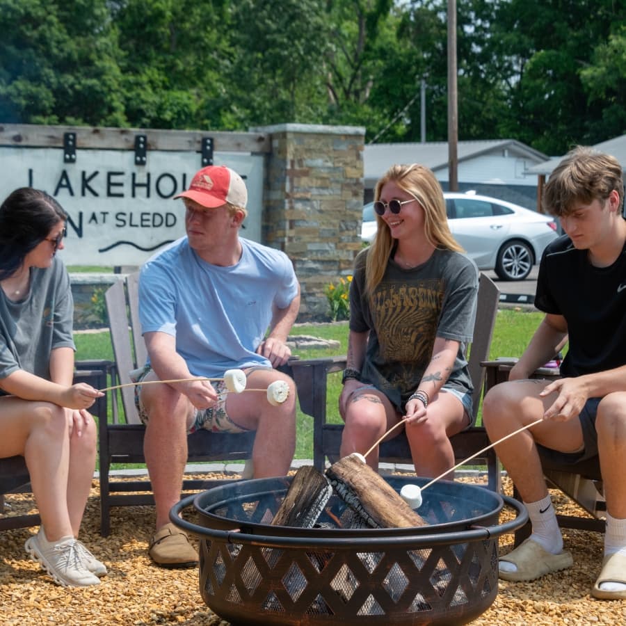 Four young people roast marshmallows around a fire pit outside a lakehouse.