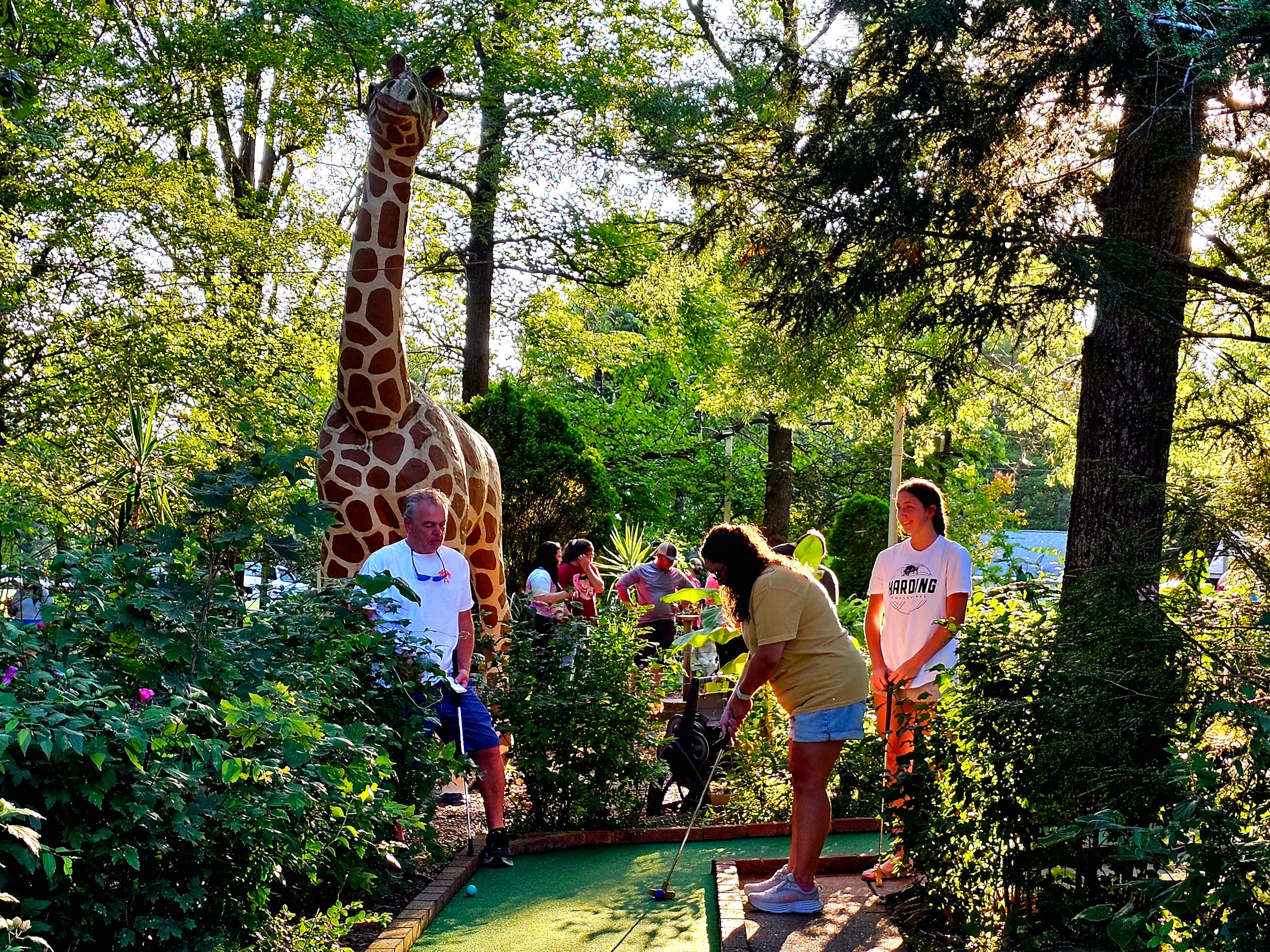 A mini-golf scene featuring players along with a large statue of a giraffe surrounded by lush greenery. A mini-golf scene featuring players along with a large statue of a giraffe surrounded by lush greenery.
