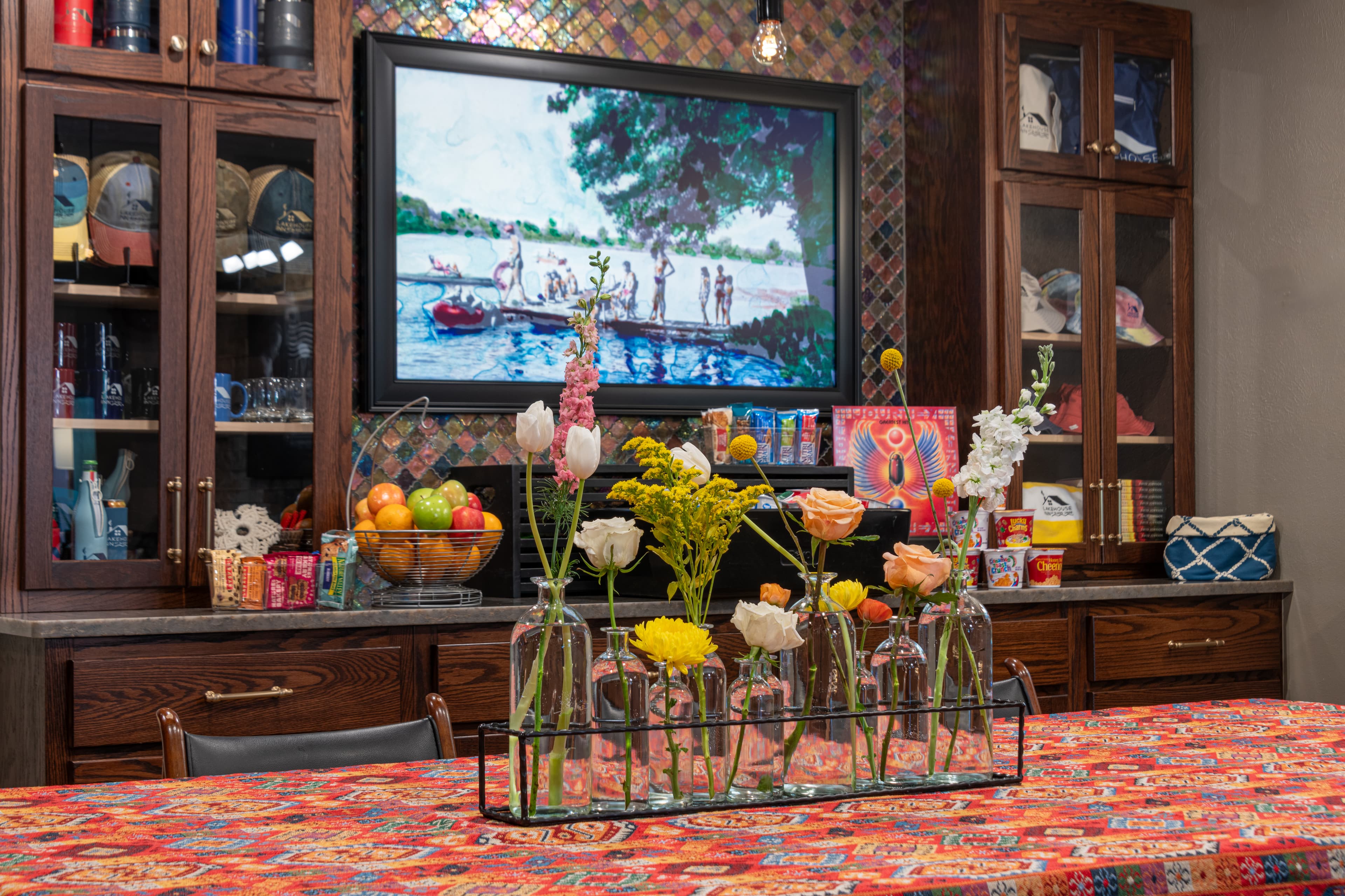 A colorful dining table adorned with flowers and surrounded by wooden cabinets displaying various items, alongside a vibrant painting of a lakeside scene.