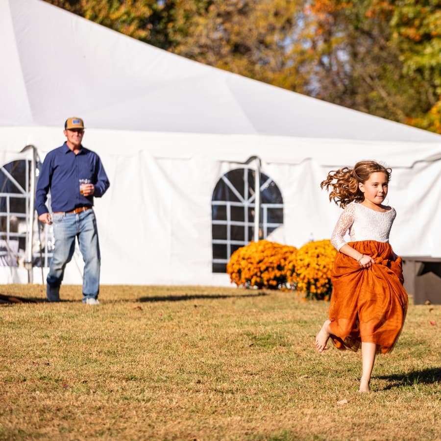 A young girl in an orange skirt runs joyfully across a grassy area while a man stands nearby holding a drink.