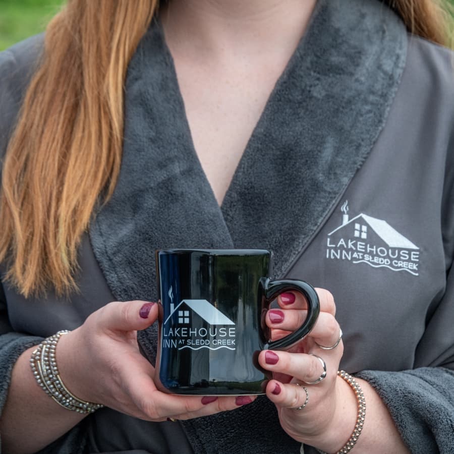 A person in a gray robe holds a black mug with the "Lakehouse Inn at Sledd Creek" logo.