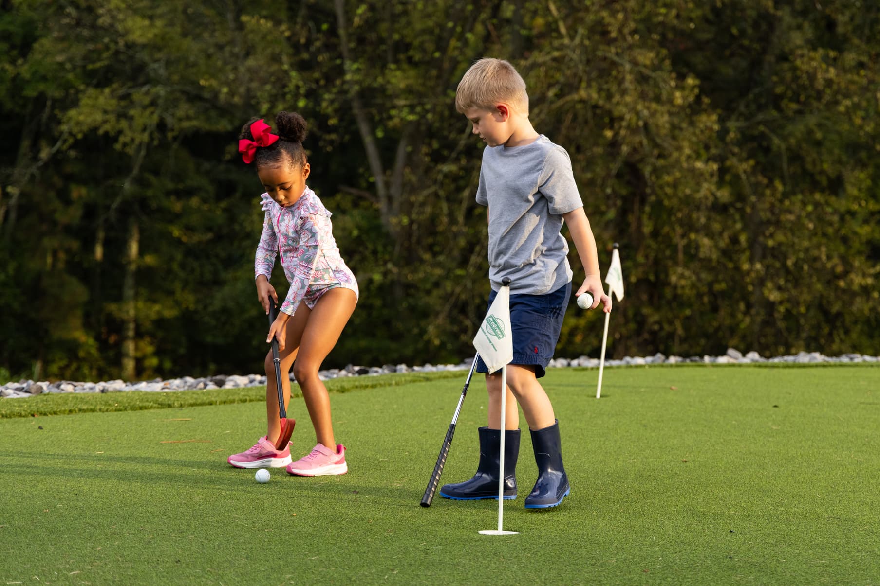 Two children playing mini-golf on a green, one preparing to putt and the other holding a ball.