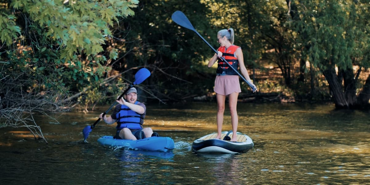 A man kayaking and a woman paddleboarding on a calm river surrounded by trees.