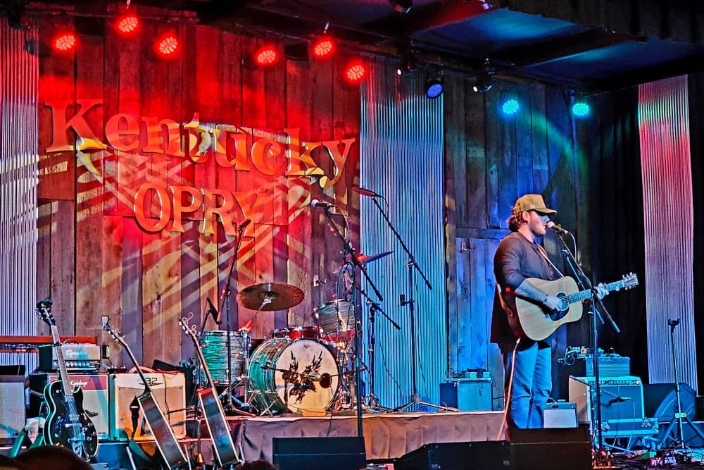 A musician performs on stage at the Kentucky Opry, with colorful lights and instruments in the background.