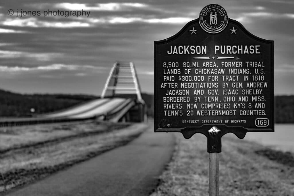 A historical marker titled "Jackson Purchase" outlines the area's history and borders, set against a black-and-white landscape. A historical marker titled "Jackson Purchase" outlines the area's history and borders, set against a black-and-white landscape.