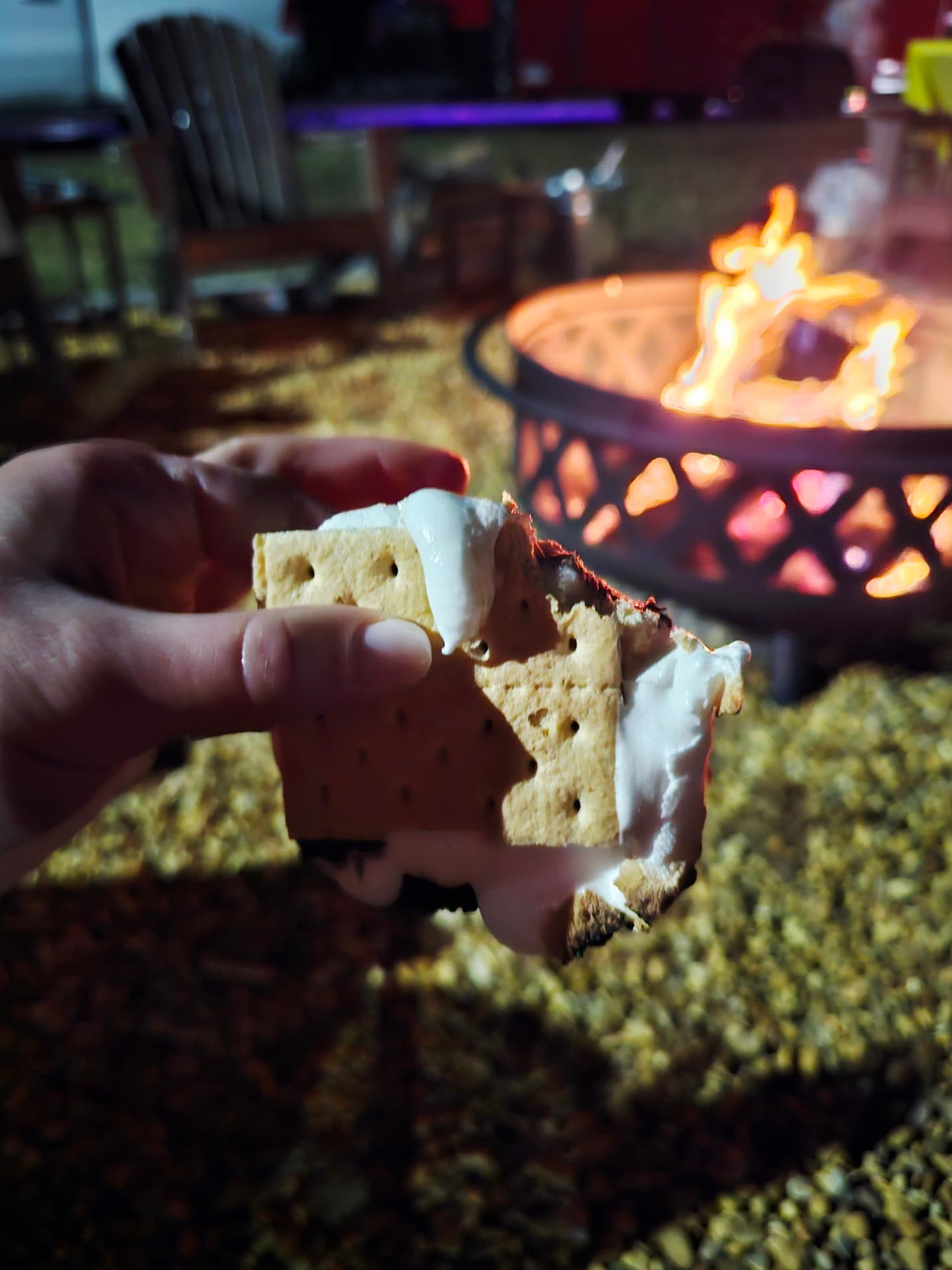 A hand holds a partially eaten s'more with melted marshmallow in front of a fire pit.