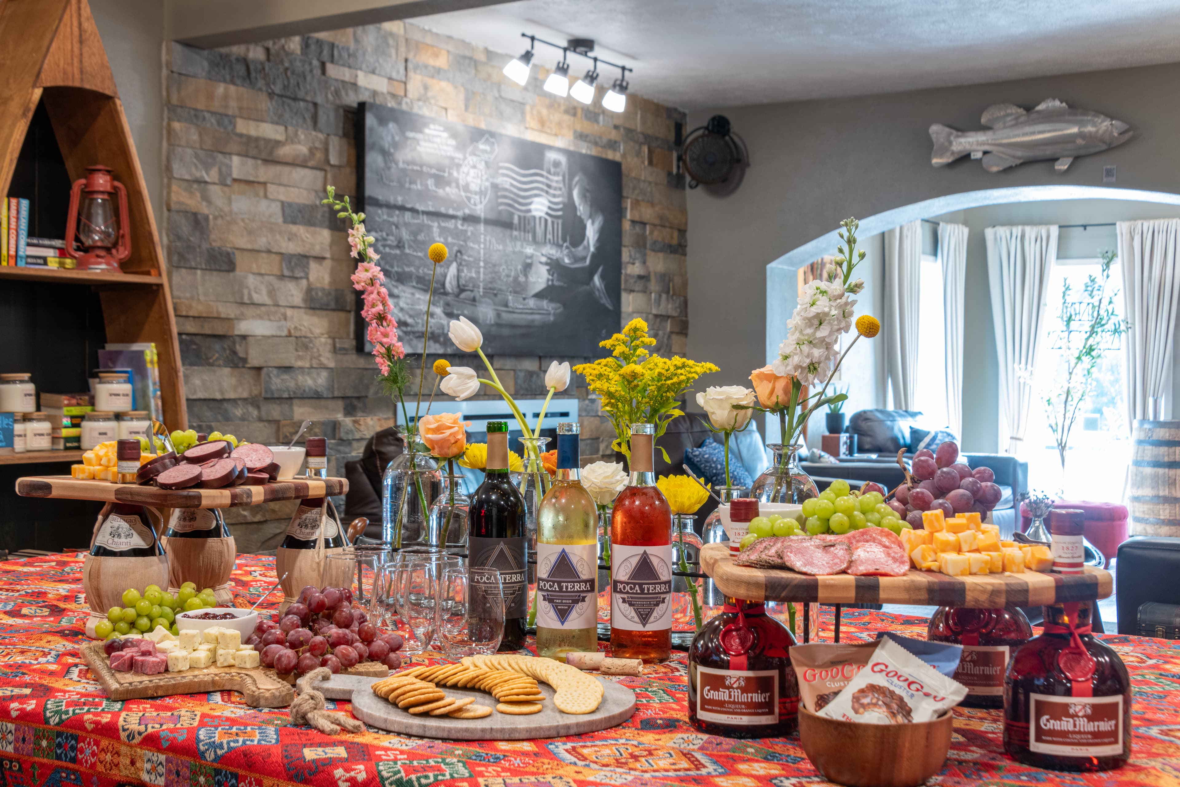 A beautifully arranged table filled with various cheeses, meats, fruits, and beverages, set against a cozy room backdrop.