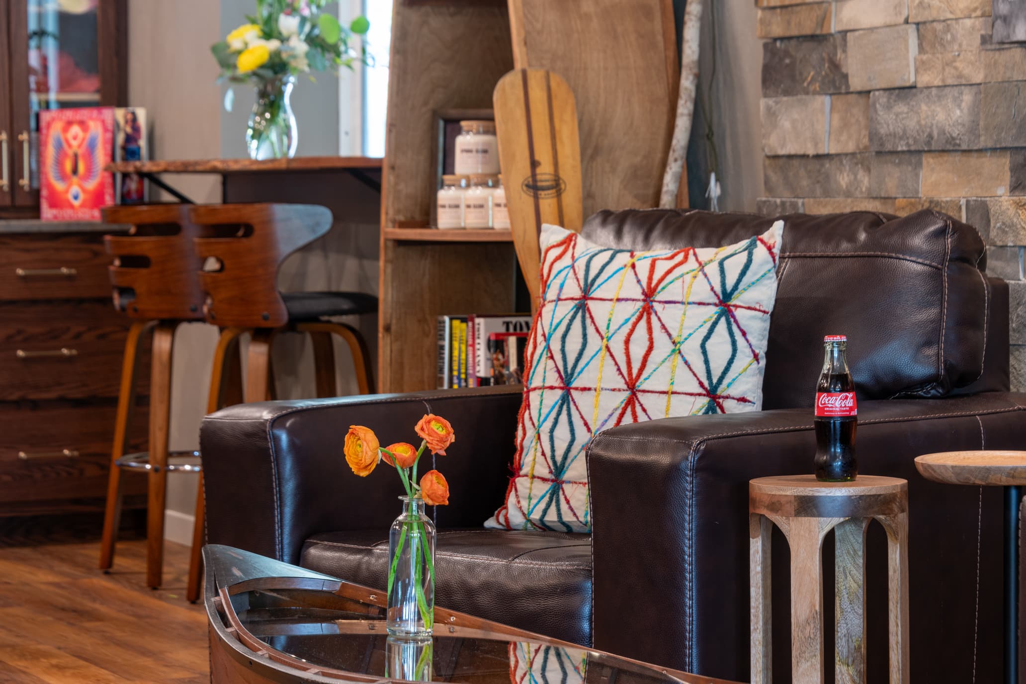 A cozy living room scene featuring a leather armchair with a colorful patterned pillow, a small vase of orange flowers, and a Coca-Cola bottle on a wooden side table.