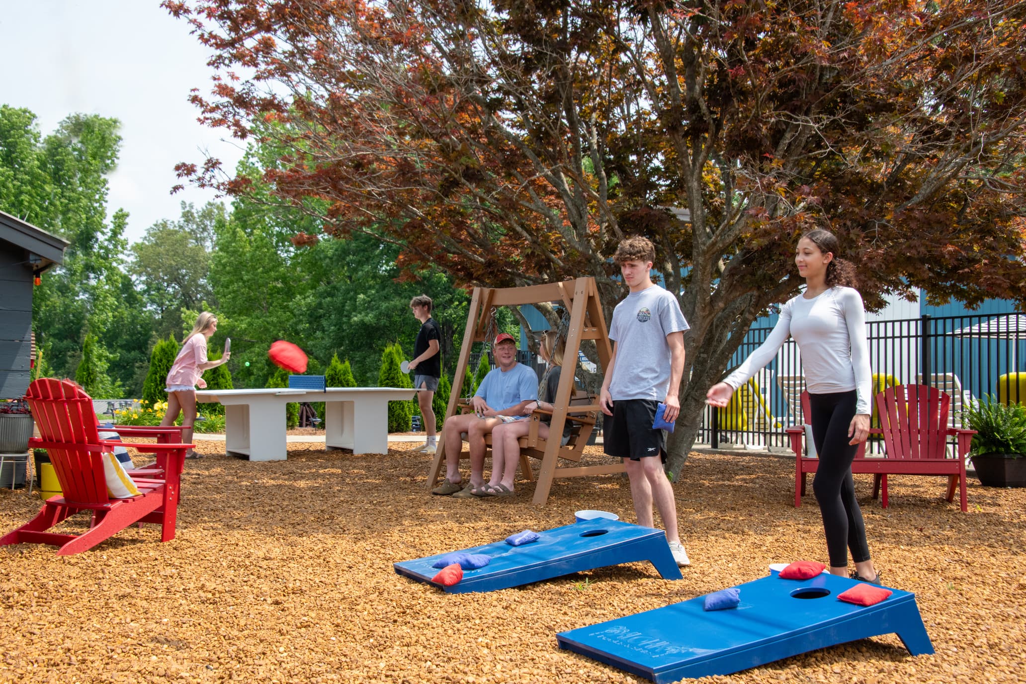 A group of six people enjoys outdoor games and relaxation in a backyard setting with seating and a shaded tree.