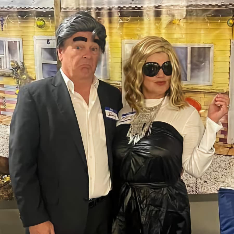 A man and woman pose together at a party, with the man wearing a suit and exaggerated hairstyle, and the woman dressed in a black dress and large sunglasses.