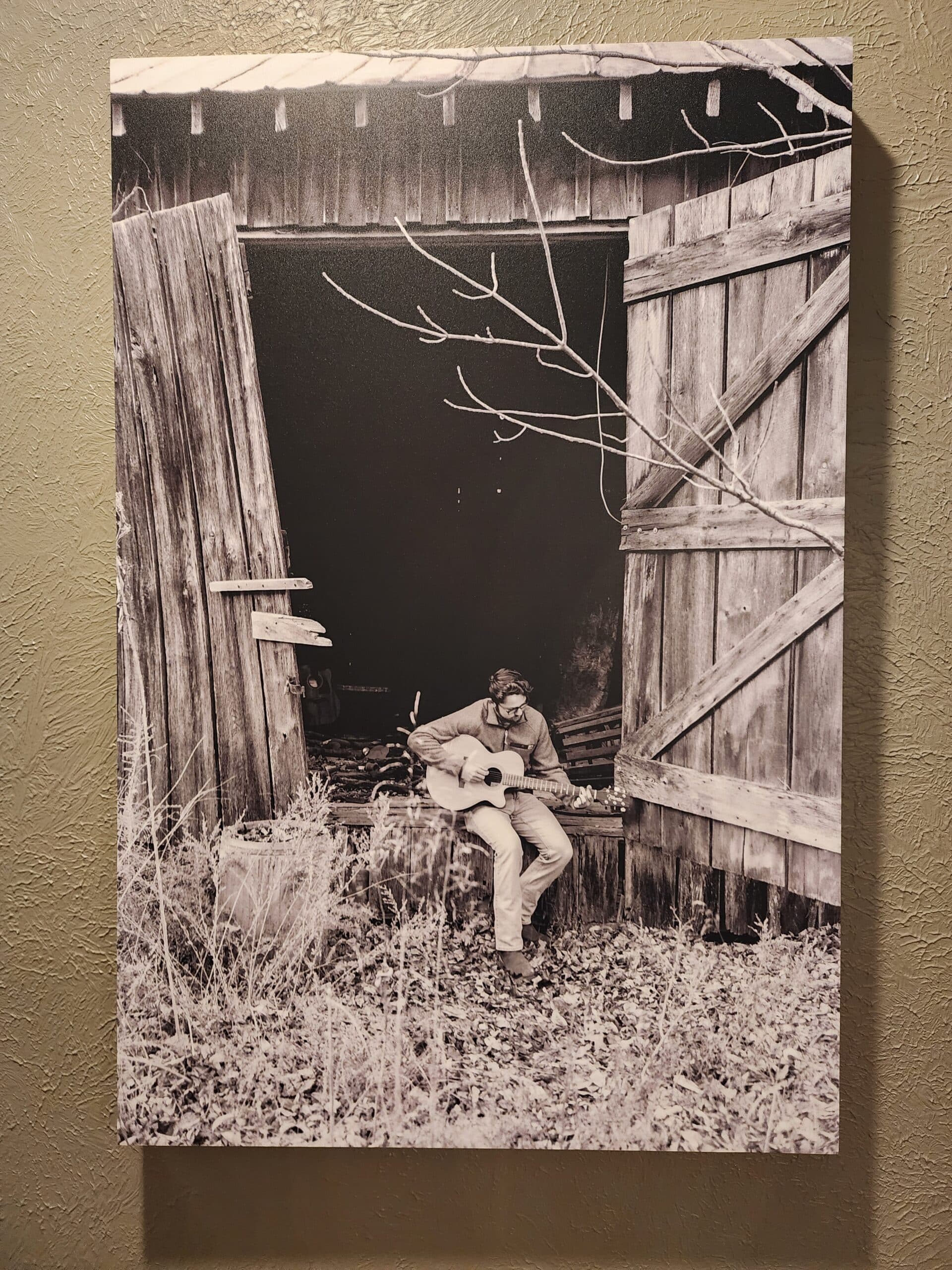 A man sits on the ground playing guitar in front of an open barn door, surrounded by rustic wooden beams and overgrown grass. A man sits on the ground playing guitar in front of an open barn door, surrounded by rustic wooden beams and overgrown grass.