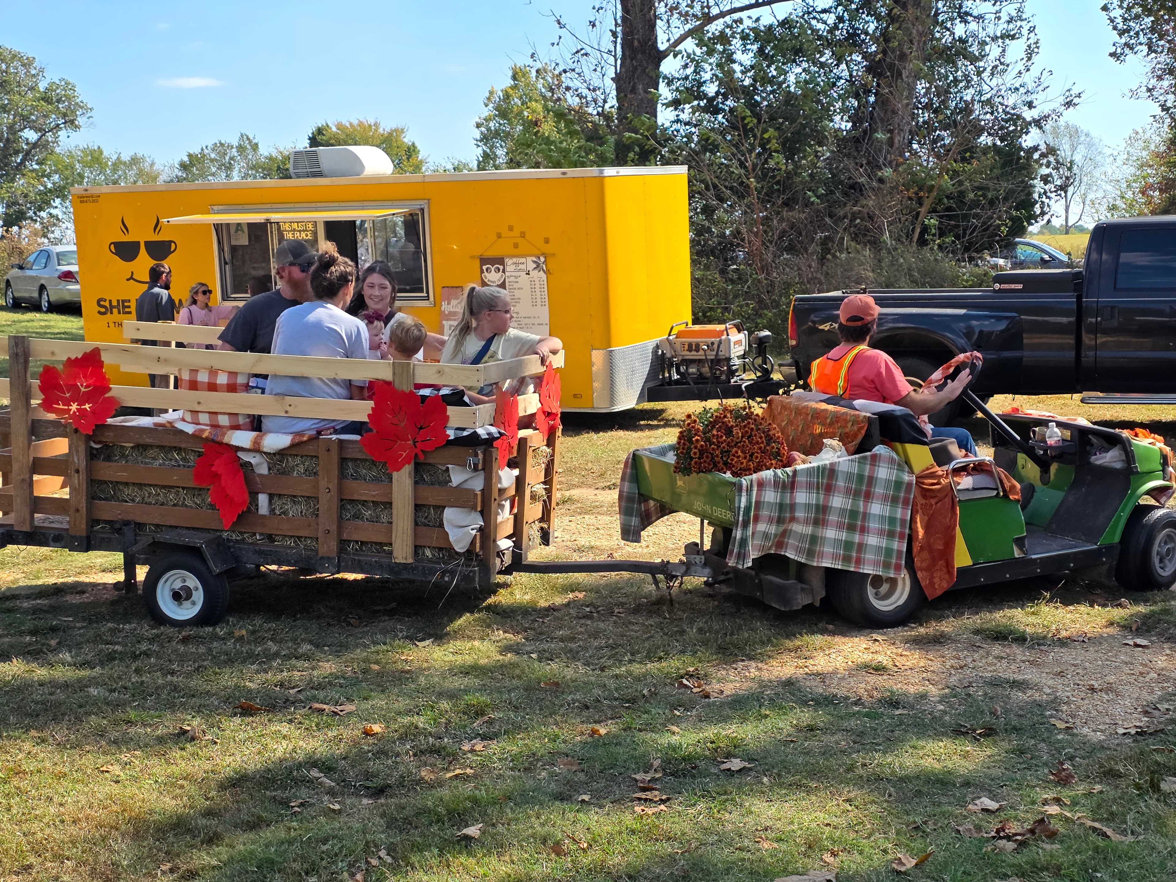 A group of people enjoys a hayride in a trailer pulled by a small vehicle, with a food truck in the background.