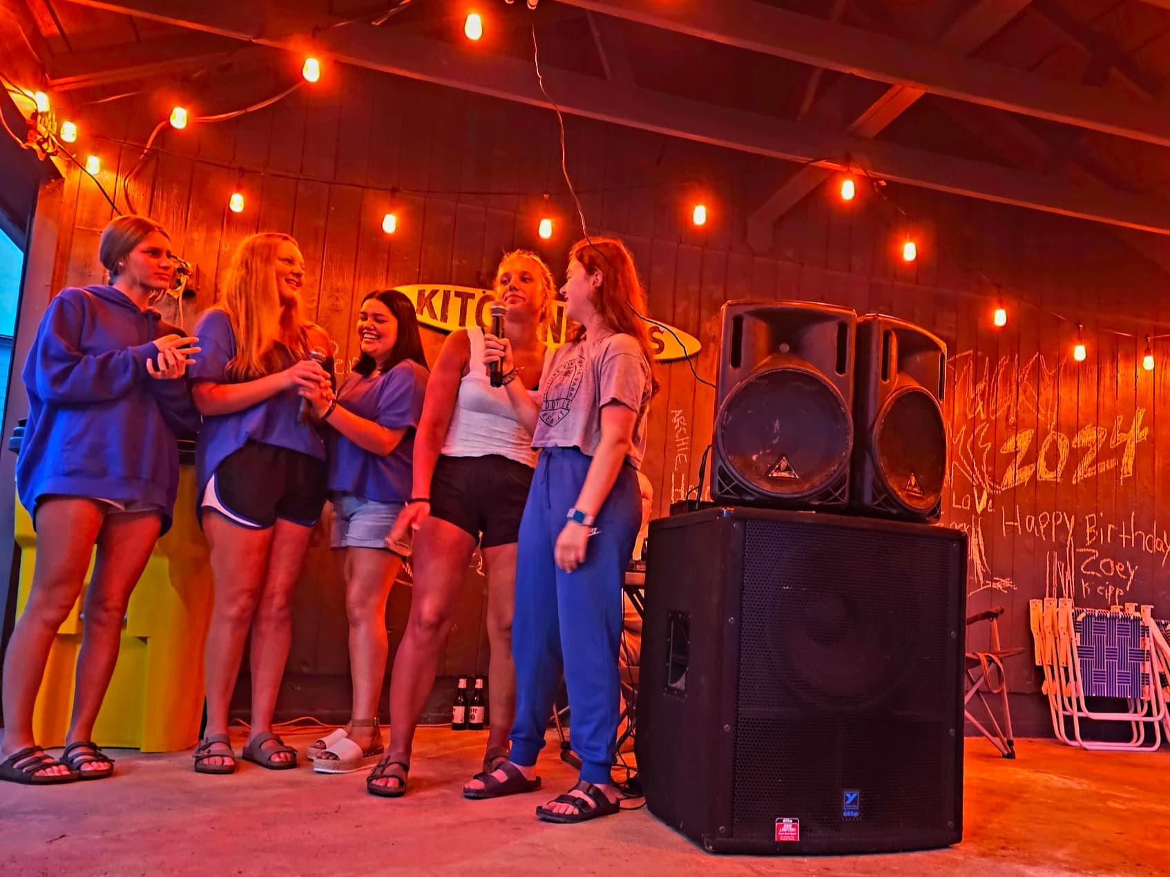 A group of five young women sing together in a warmly lit room decorated for a birthday celebration.