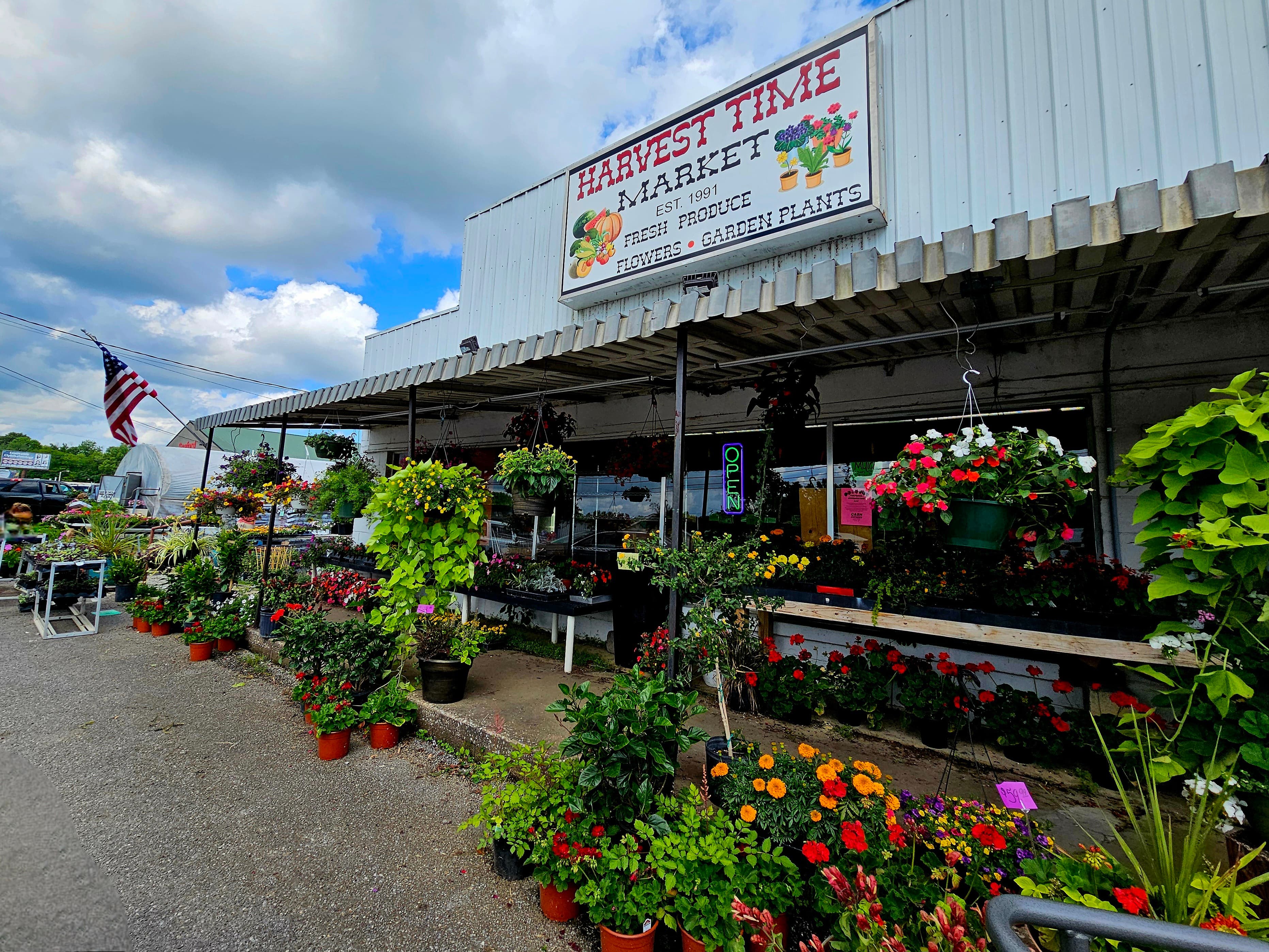 A colorful flower market with hanging plants and an American flag under a partly cloudy sky.