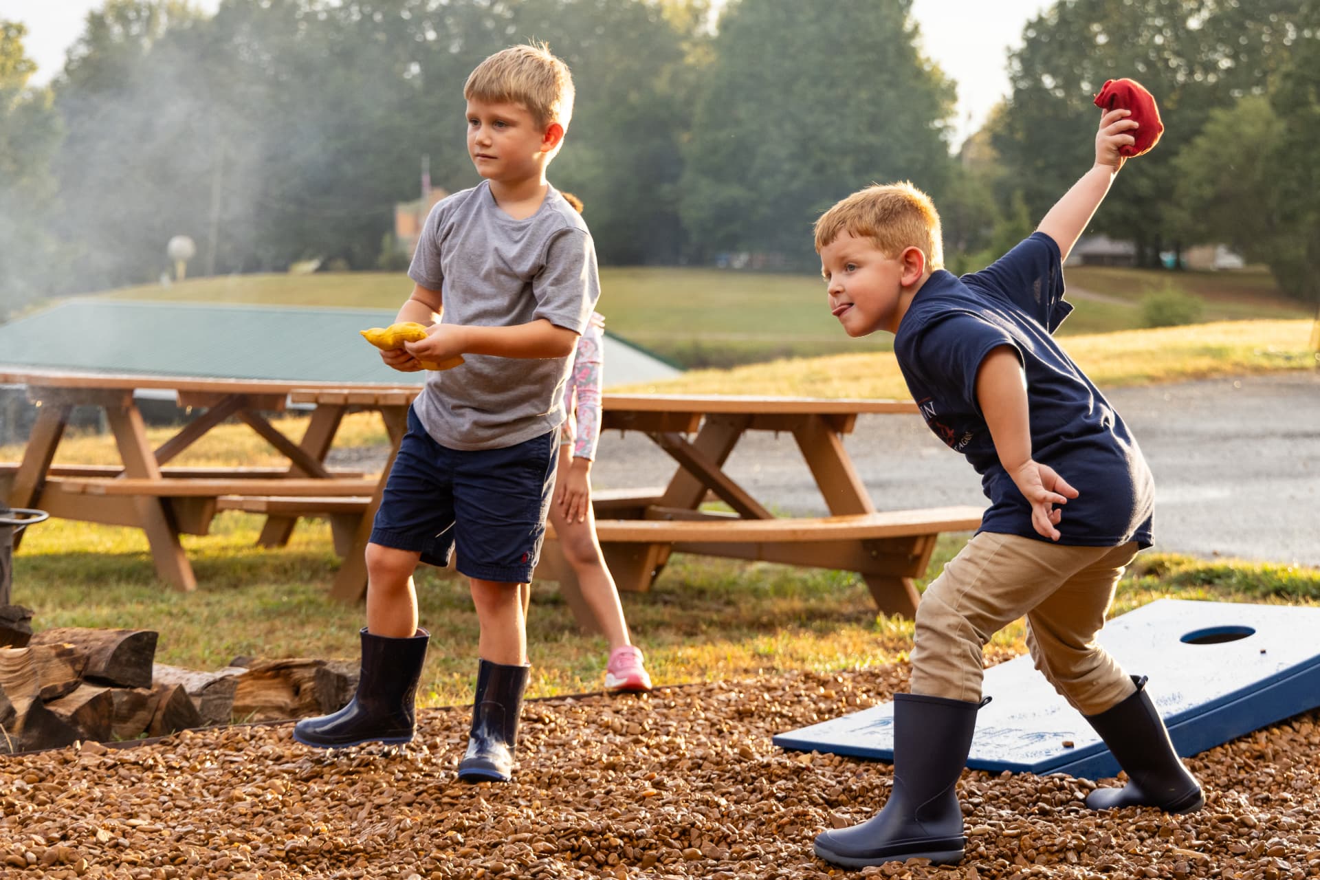 Two boys play a bean bag toss game outdoors while a third child stands nearby.