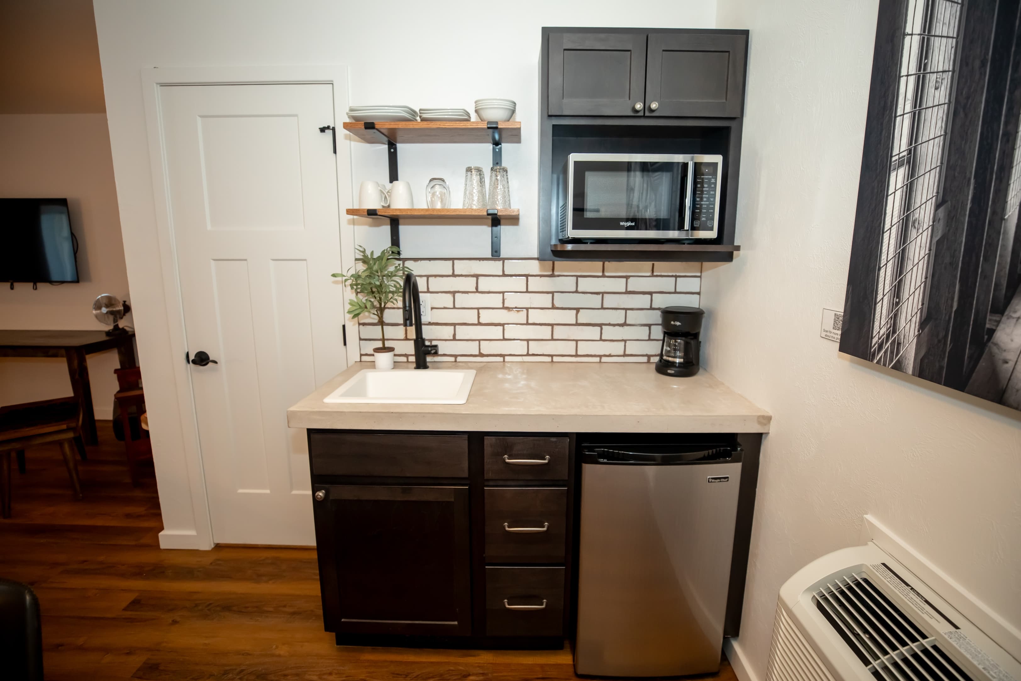 A modern kitchenette featuring a sink, microwave, refrigerator, and open shelving with dishes.