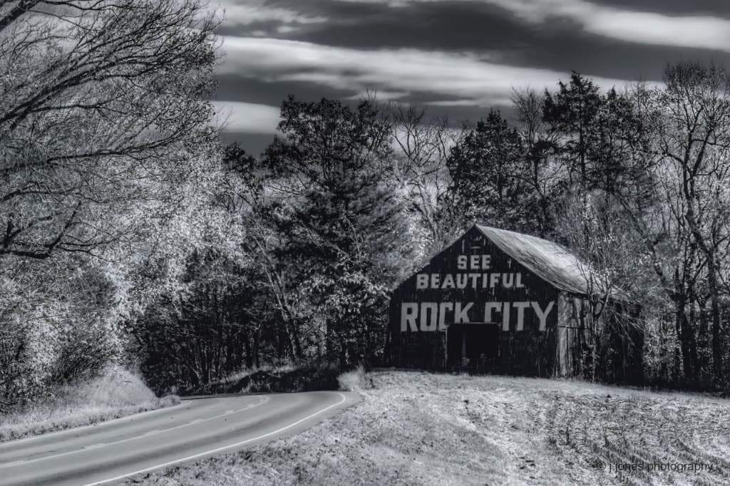 Black and white image of a rustic barn with a painted sign reading "See Beautiful Rock City," situated by a winding road and surrounded by trees. Black and white image of a rustic barn with a painted sign reading "See Beautiful Rock City," situated by a winding road and surrounded by trees.