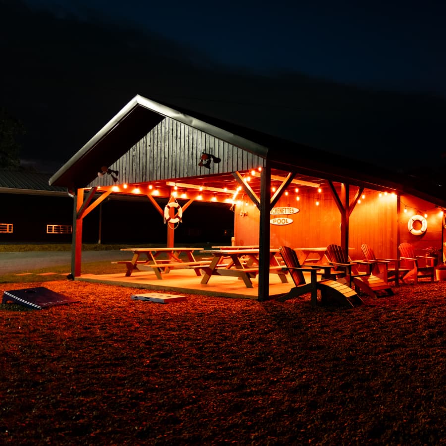 A wooden pavilion with string lights, picnic tables, and a pool sign, illuminated at night.