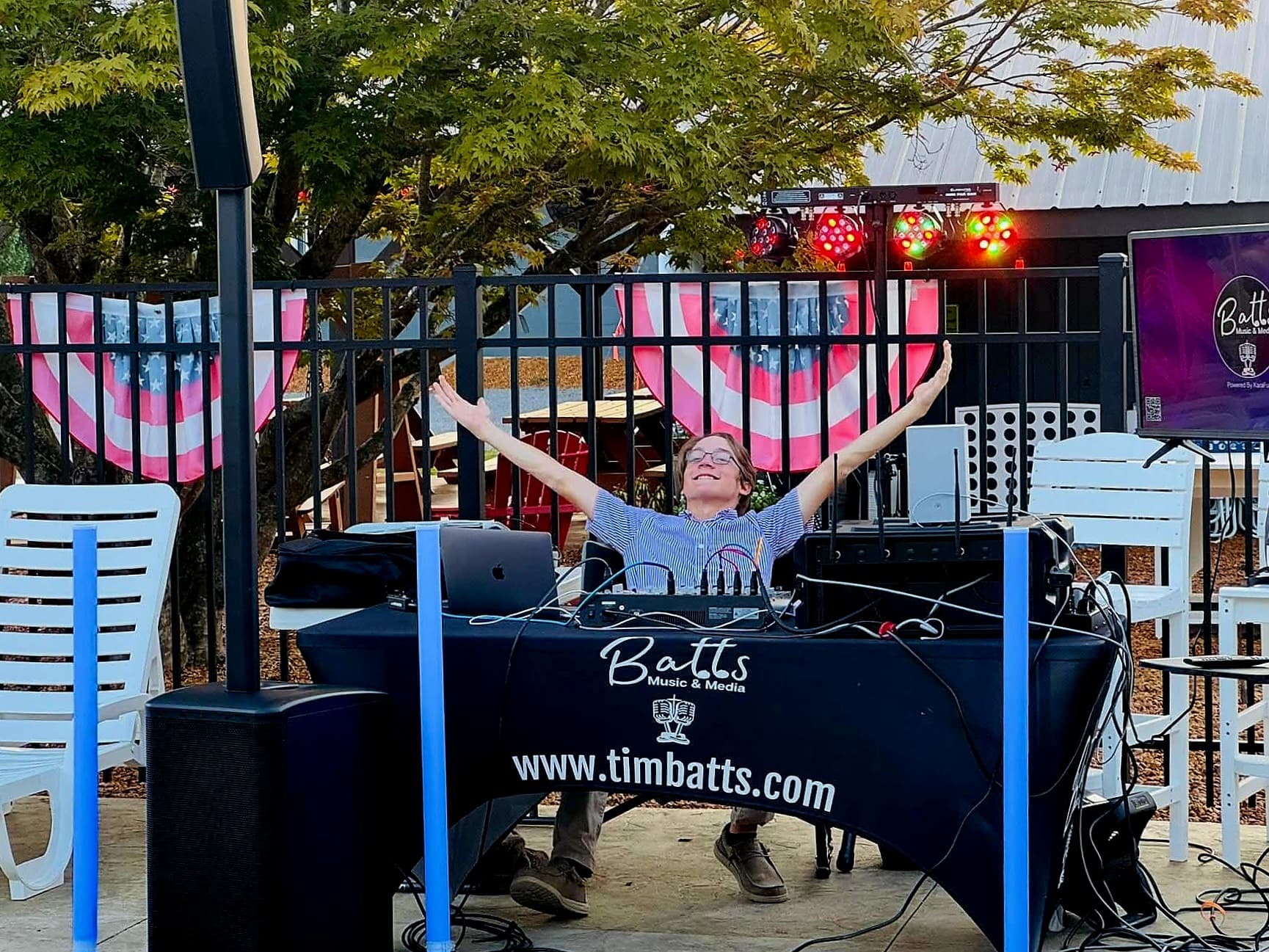 A DJ joyfully gestures while behind a soundboard at an outdoor event.