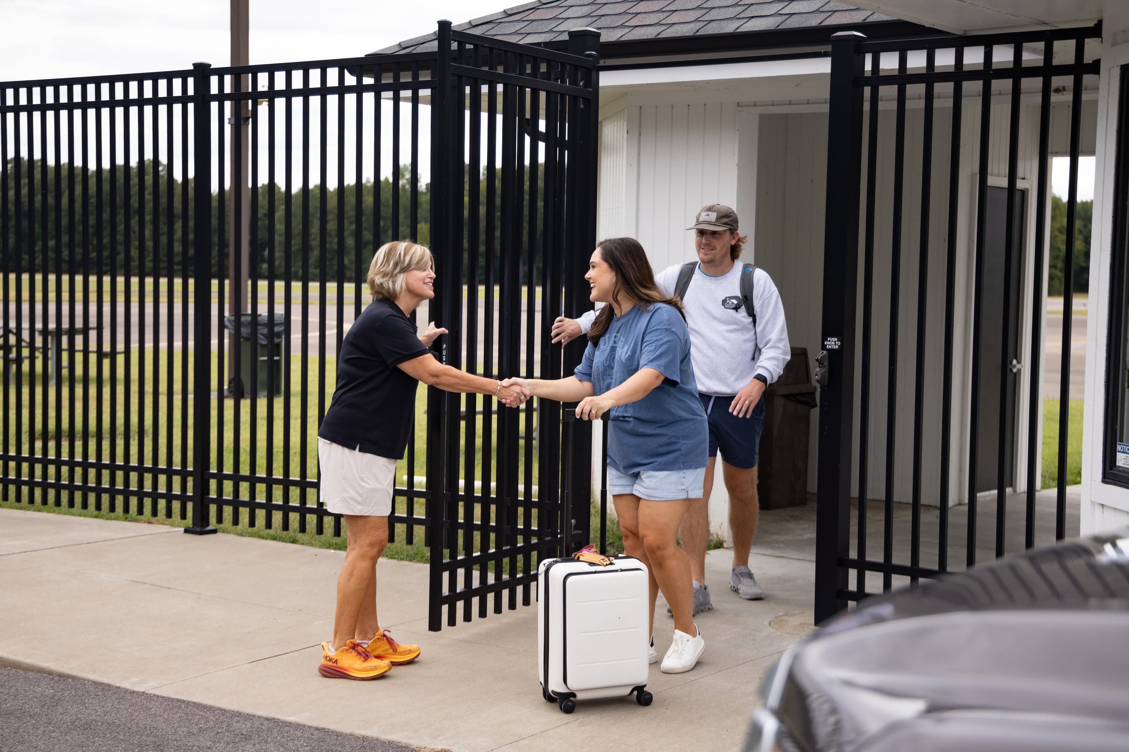 Two women shake hands at a gated entrance while a man stands behind them.