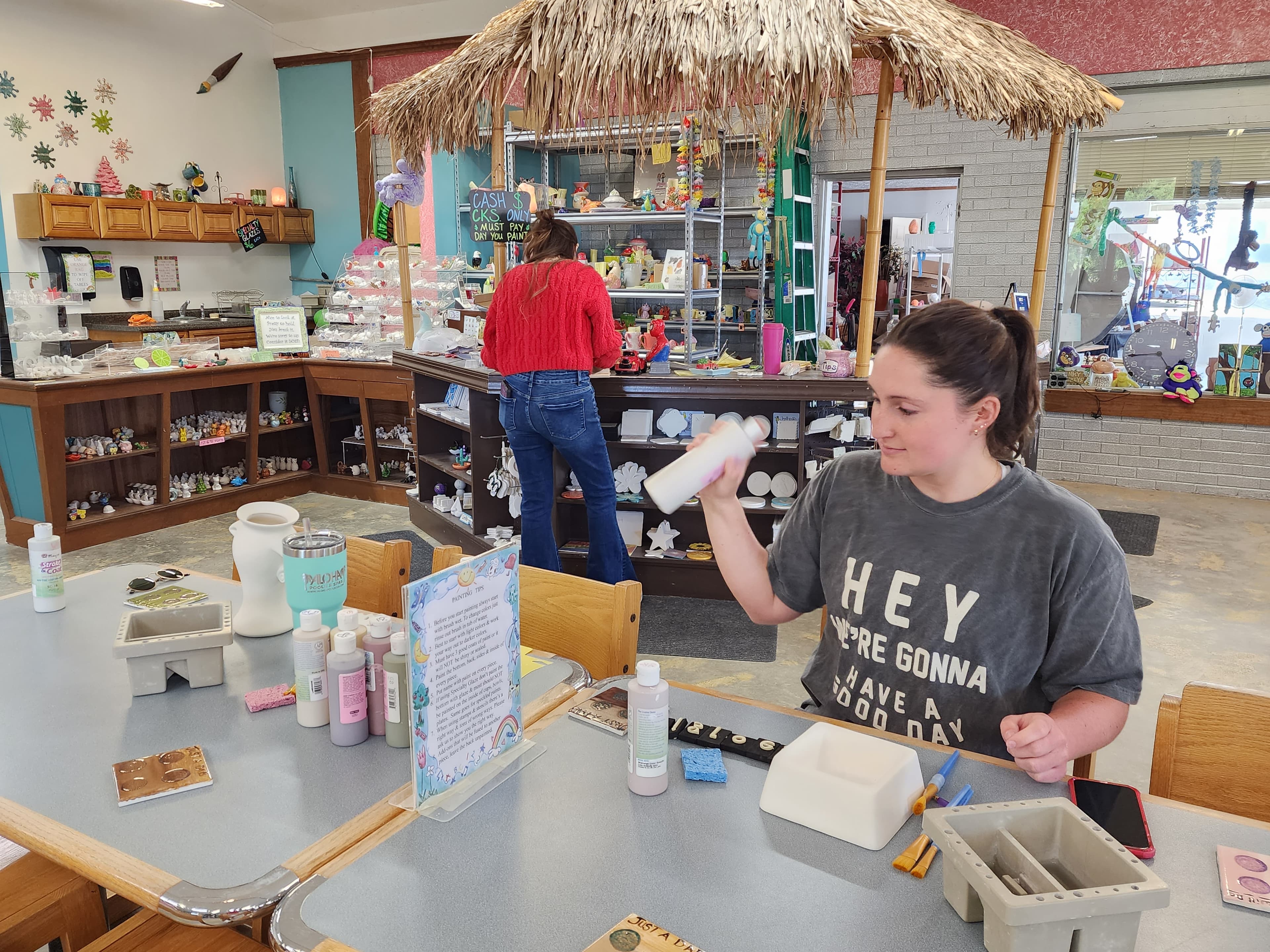A woman works on a craft project at a table filled with paint supplies, while another person browses a colorful display in a creative studio.