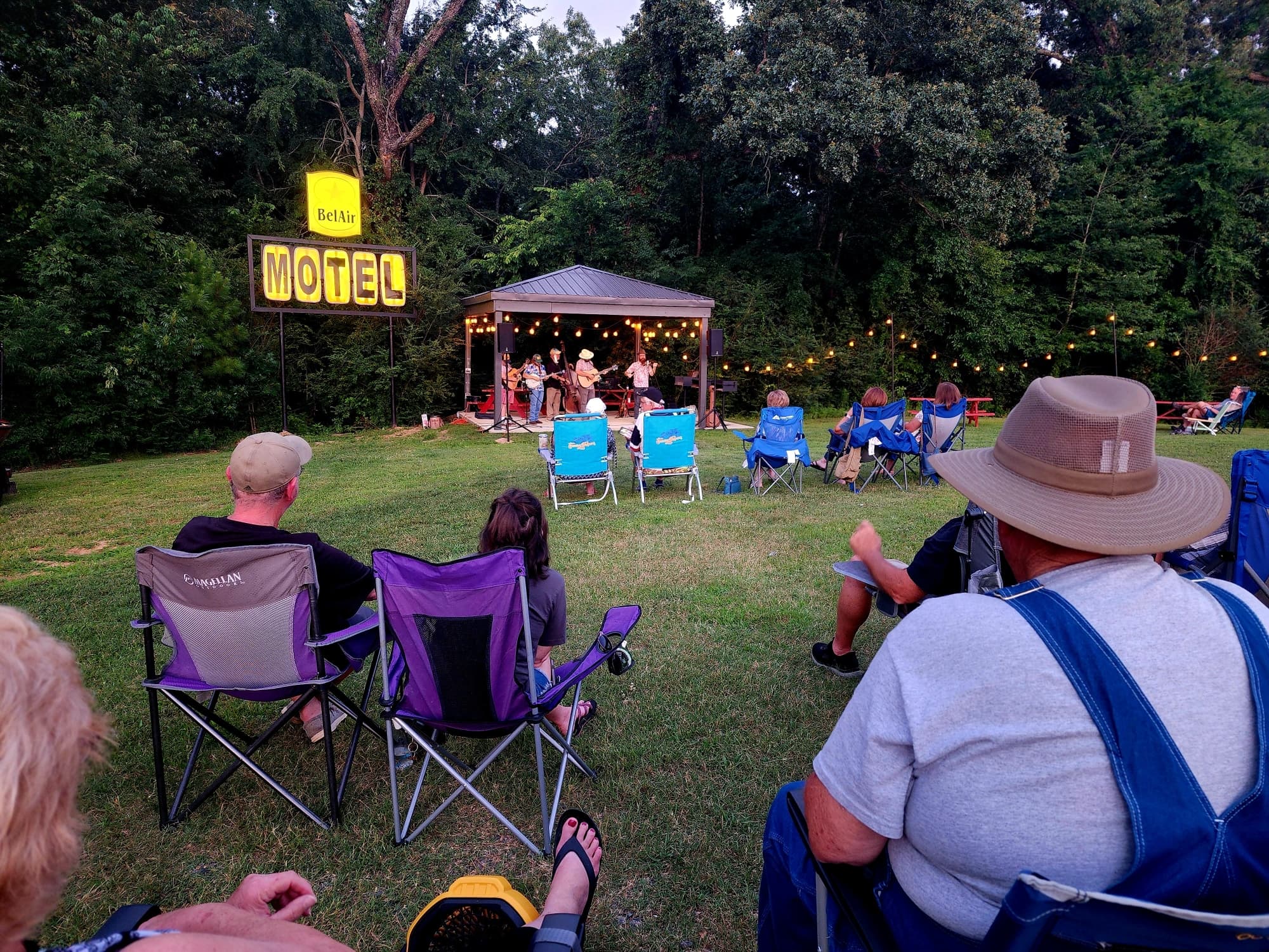 A small crowd of people sits in lawn chairs enjoying a live music performance near a vintage motel sign.