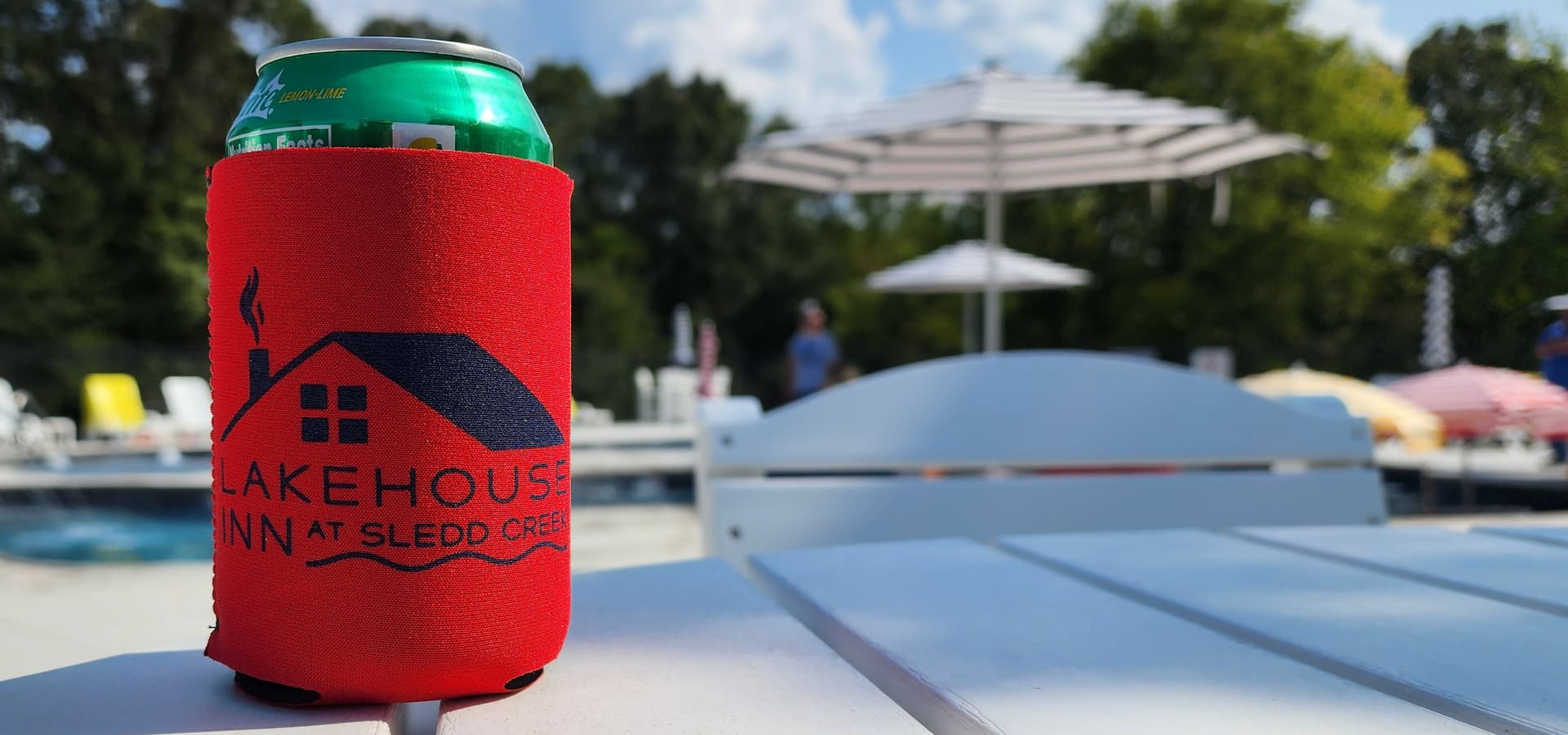 A red koozie with a logo sits on a white table beside a pool on a sunny day.