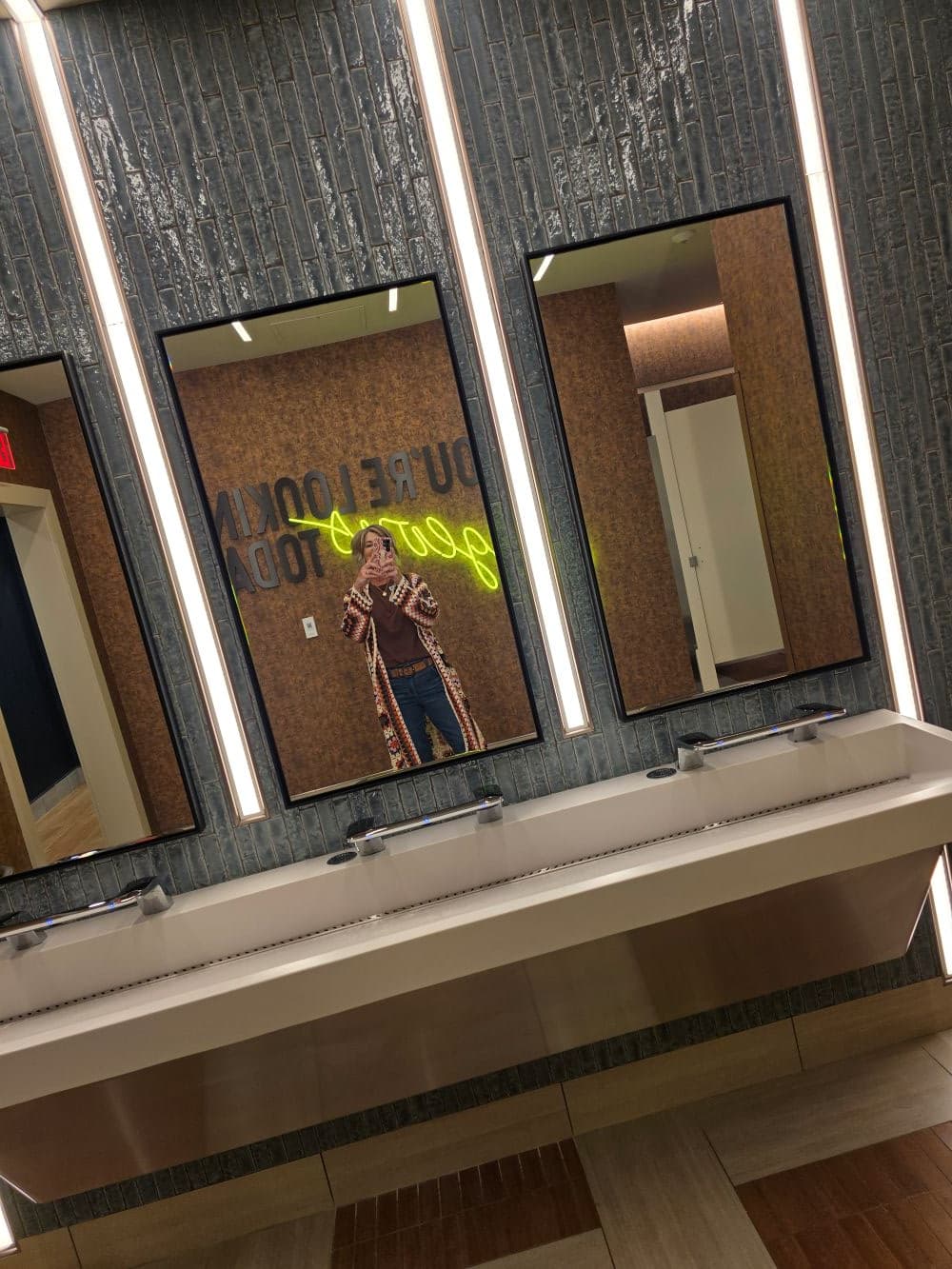 A person stands in front of three mirrors in a stylish restroom with a neon sign on the wall.