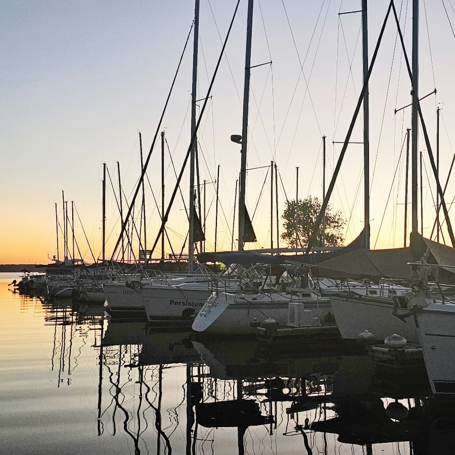 Sailboats lined up at a marina during sunset, reflecting on calm waters.