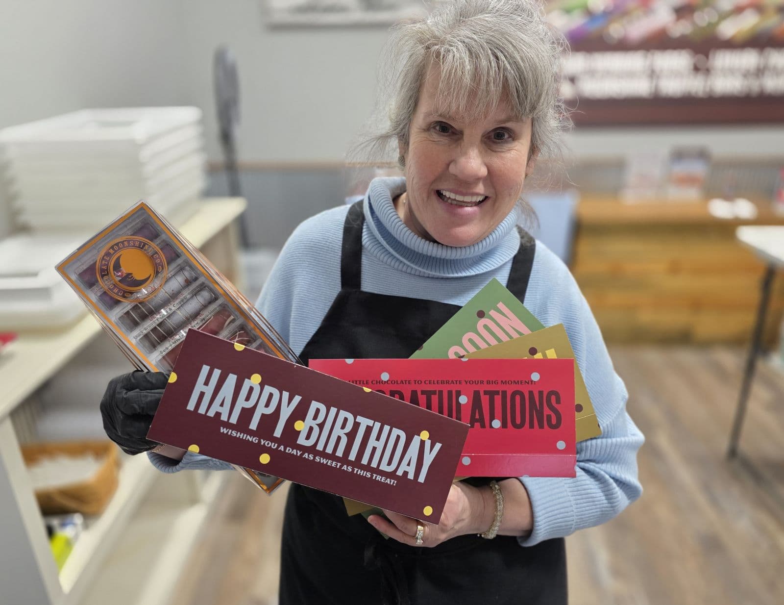A woman in an apron smiles while holding colorful signs that read "Happy Birthday" and "Congratulations."