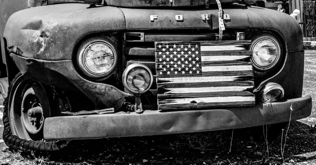 A weathered Ford truck with a distressed American flag grille sits in a gravel lot. A weathered Ford truck with a distressed American flag grille sits in a gravel lot.