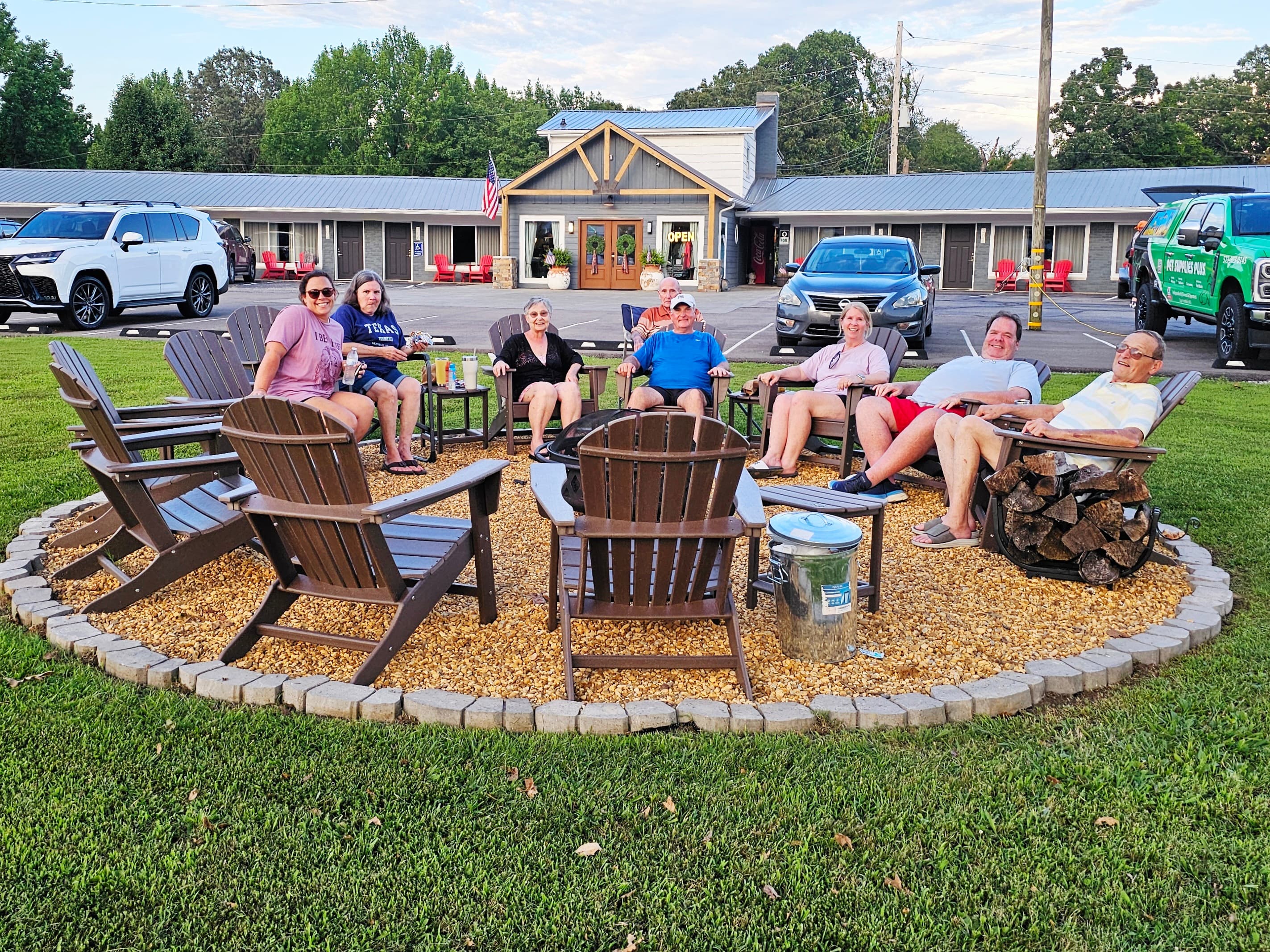 A group of eight people sits in a circular arrangement of chairs around a fire pit in a grassy area.