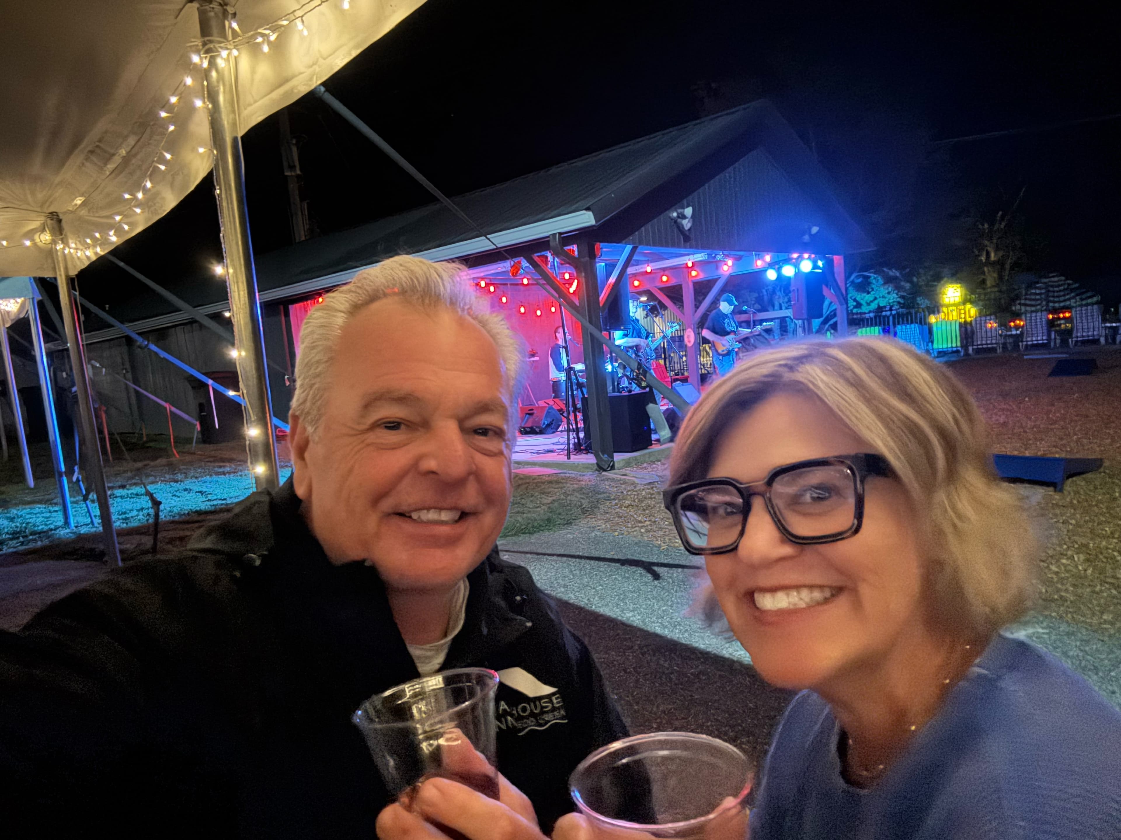 Two smiling individuals hold cups in front of a lit outdoor stage during a night event.