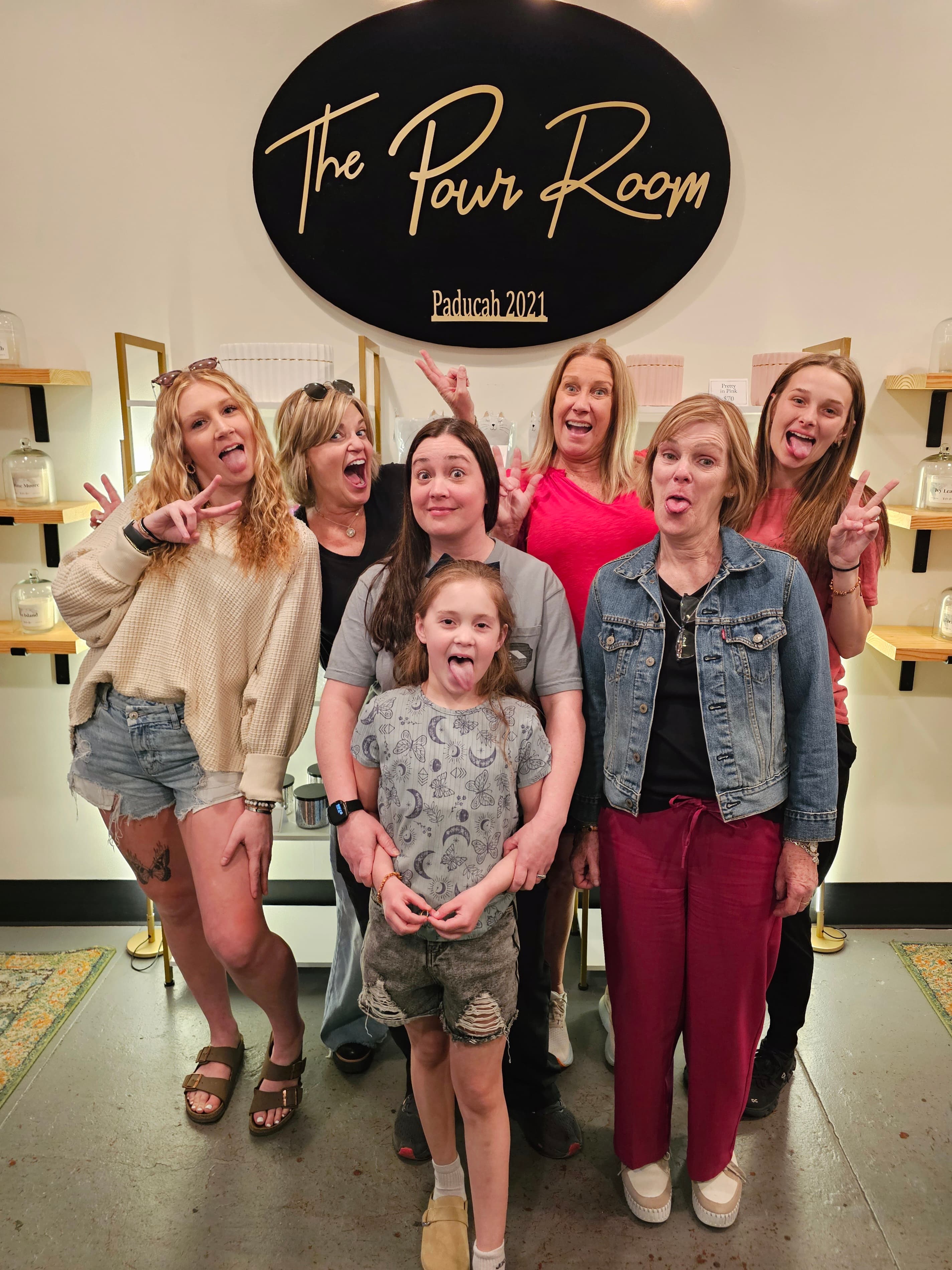 A group of seven women and a girl pose playfully in front of "The Pour Room" sign, making silly faces.