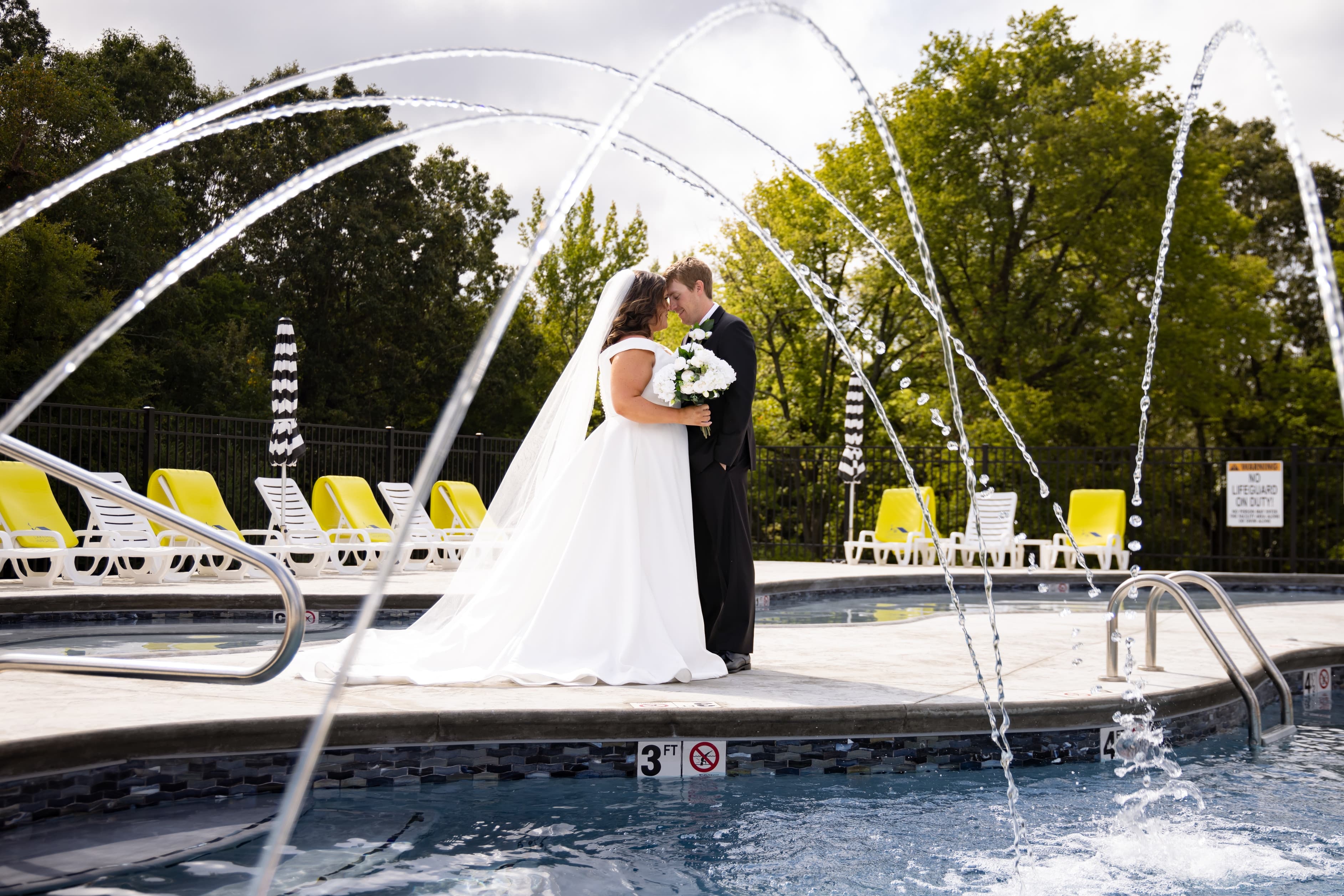 A bride and groom kiss under a fountain beside a swimming pool.