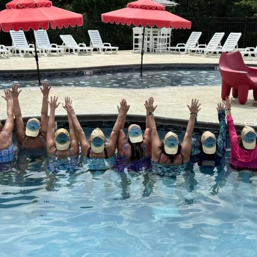 A group of women in swimsuits and hats are playfully splashing in a pool, raising their arms in celebration.