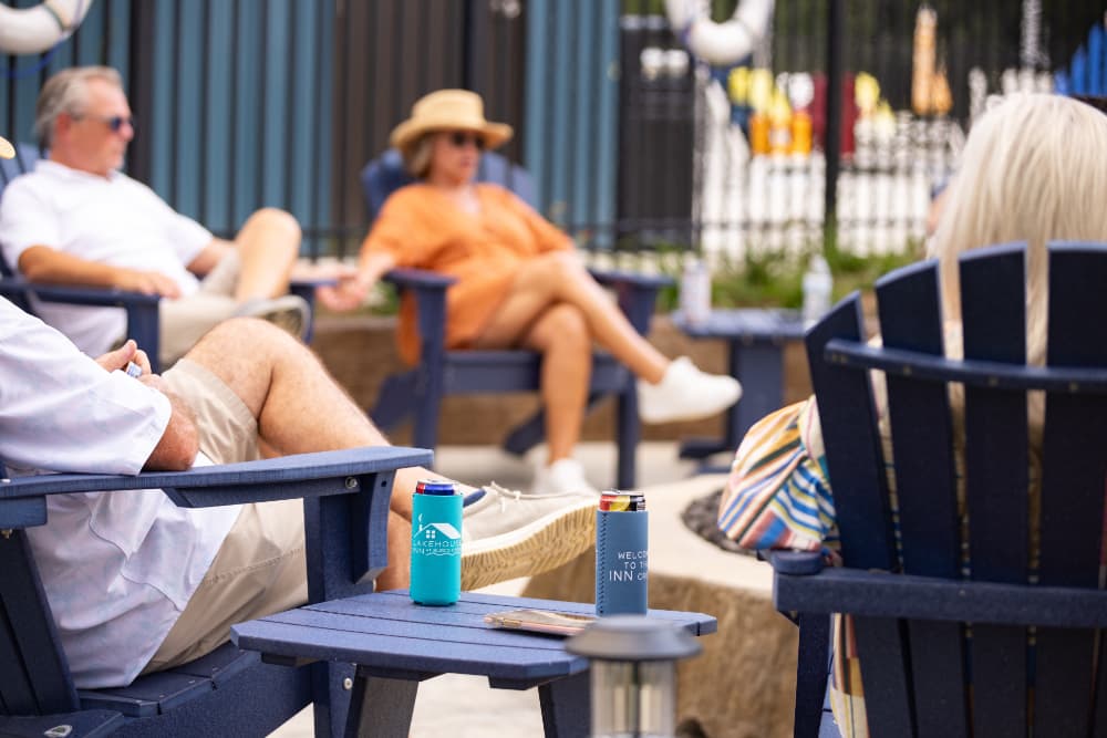 A group of people relax in outdoor chairs with drinks, enjoying a sunny day by a pool.