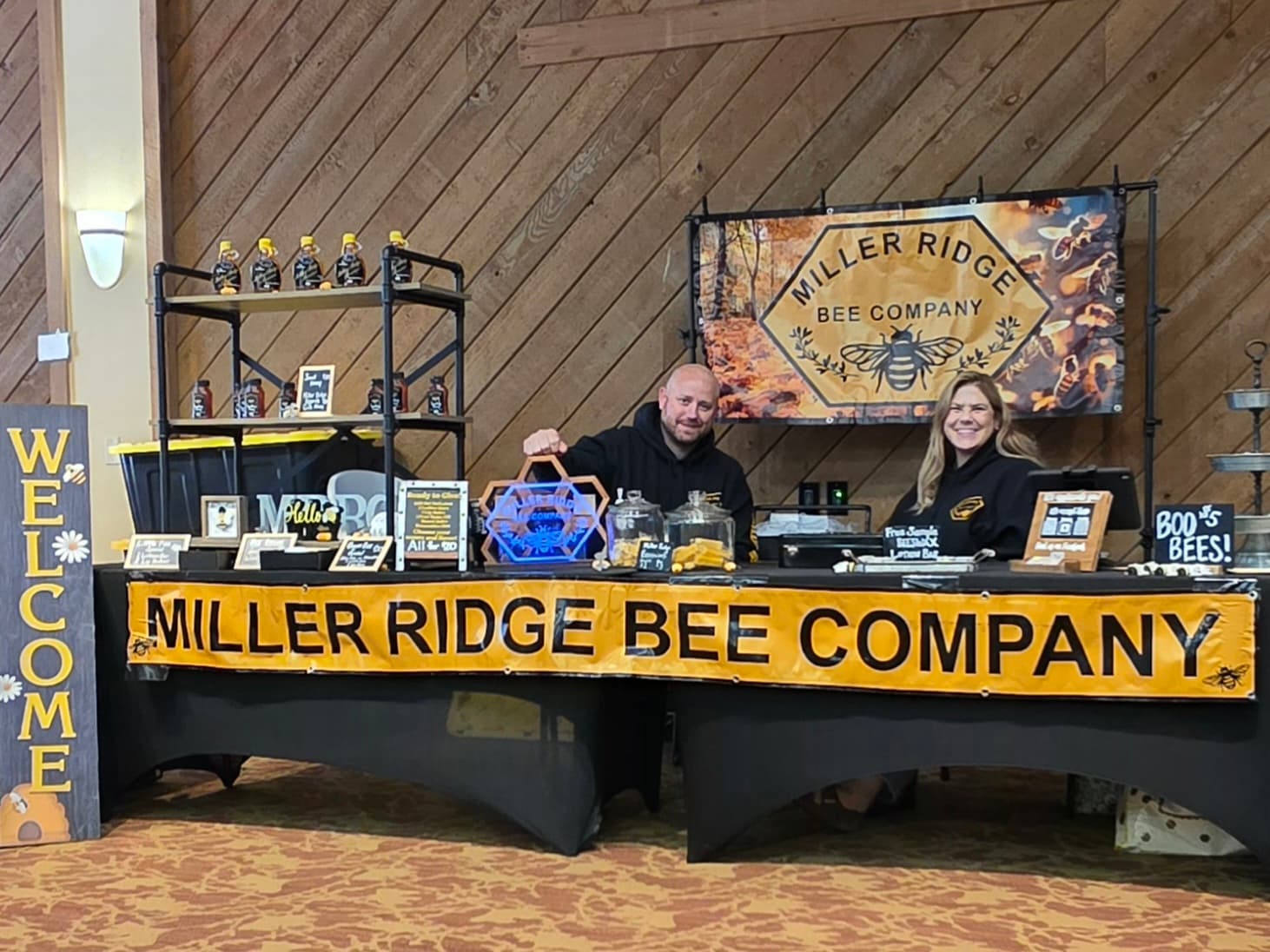 A man and woman stand behind a vendor table displaying honey products and promotional materials for Miller Ridge Bee Company.