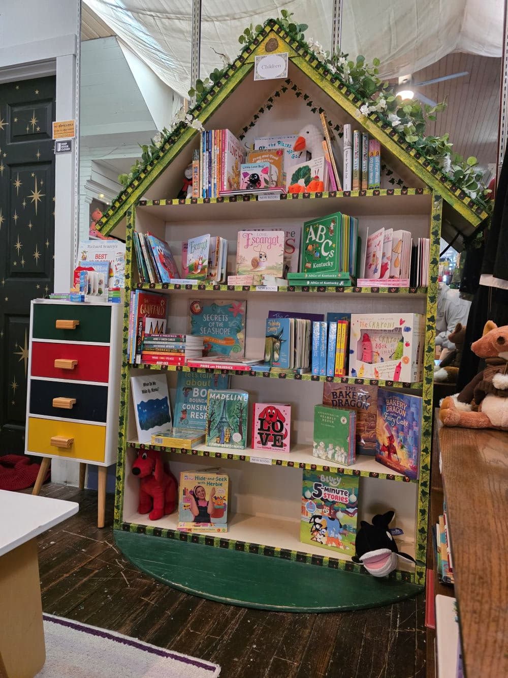 A whimsical bookshelf shaped like a house, filled with colorful children's books and adorned with decorative greenery.