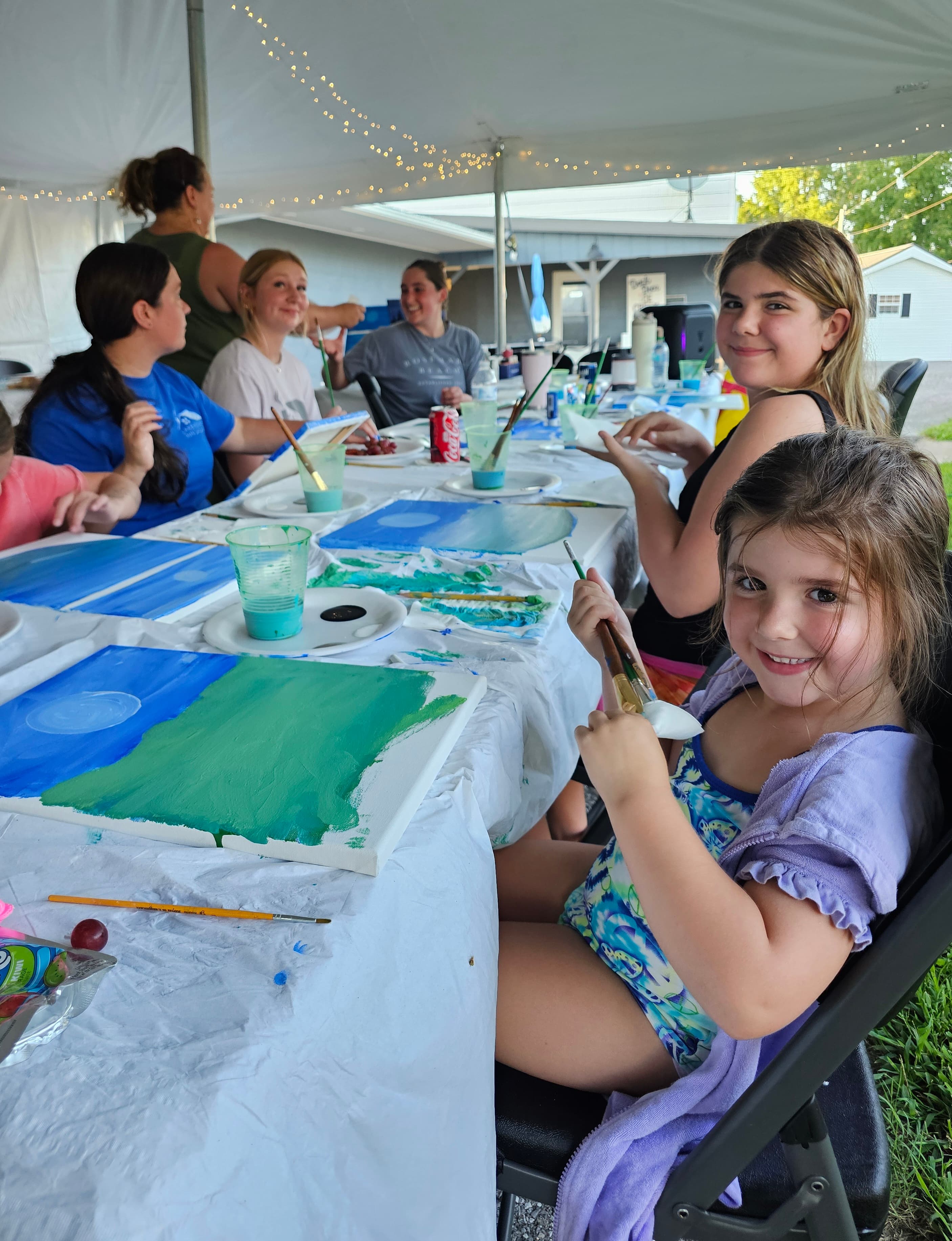 A group of people, including a young girl, enjoy a painting activity outdoors under a tent.