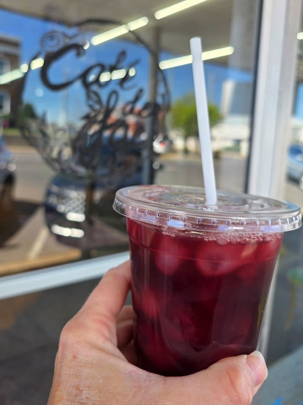 A hand holding a clear cup filled with dark red drink, in front of a café window.