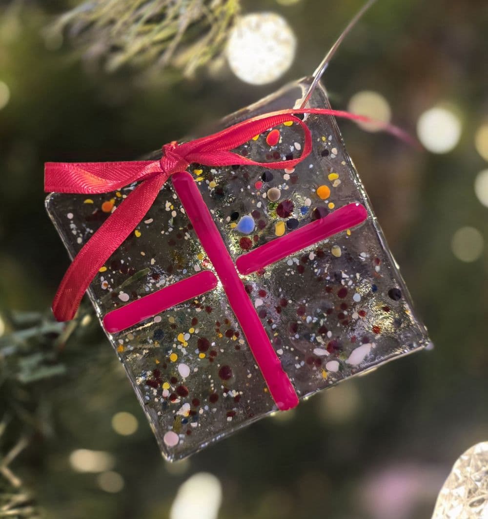 A colorful, sparkly gift ornament with a red ribbon hangs on a Christmas tree.