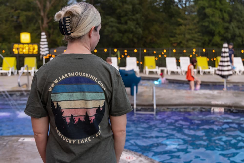A person with a ponytail wears a Lake House Inn t-shirt while standing near a pool, observing others enjoying the water.