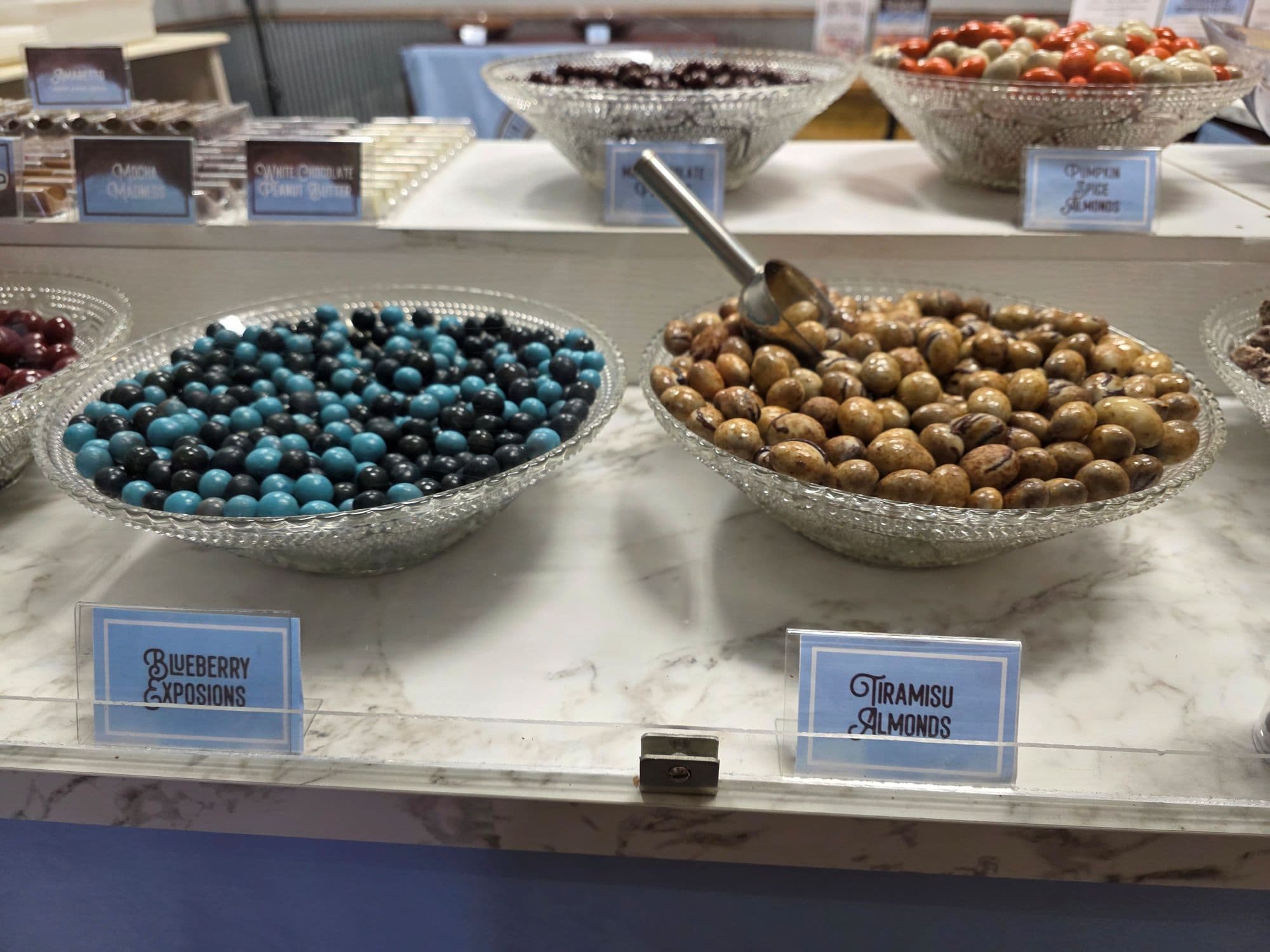 A display of colorful candy bowls labeled "Blueberry Explosions" and "Tiramisu Almonds."