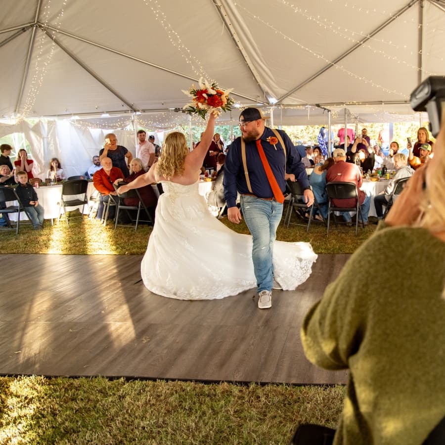 A bride in a white dress joyfully twirls with a bouquet, accompanied by a man in casual attire, on a dance floor surrounded by wedding guests.