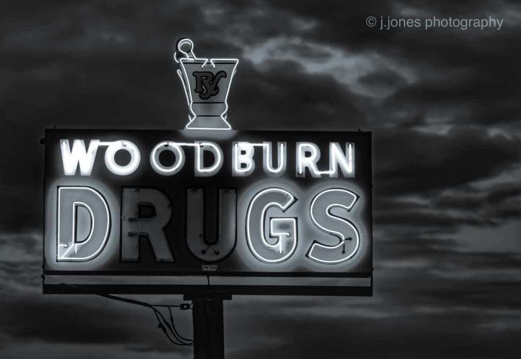 A vintage neon sign reading "Woodburn Drugs" against a cloudy sky. A vintage neon sign reading "Woodburn Drugs" against a cloudy sky.