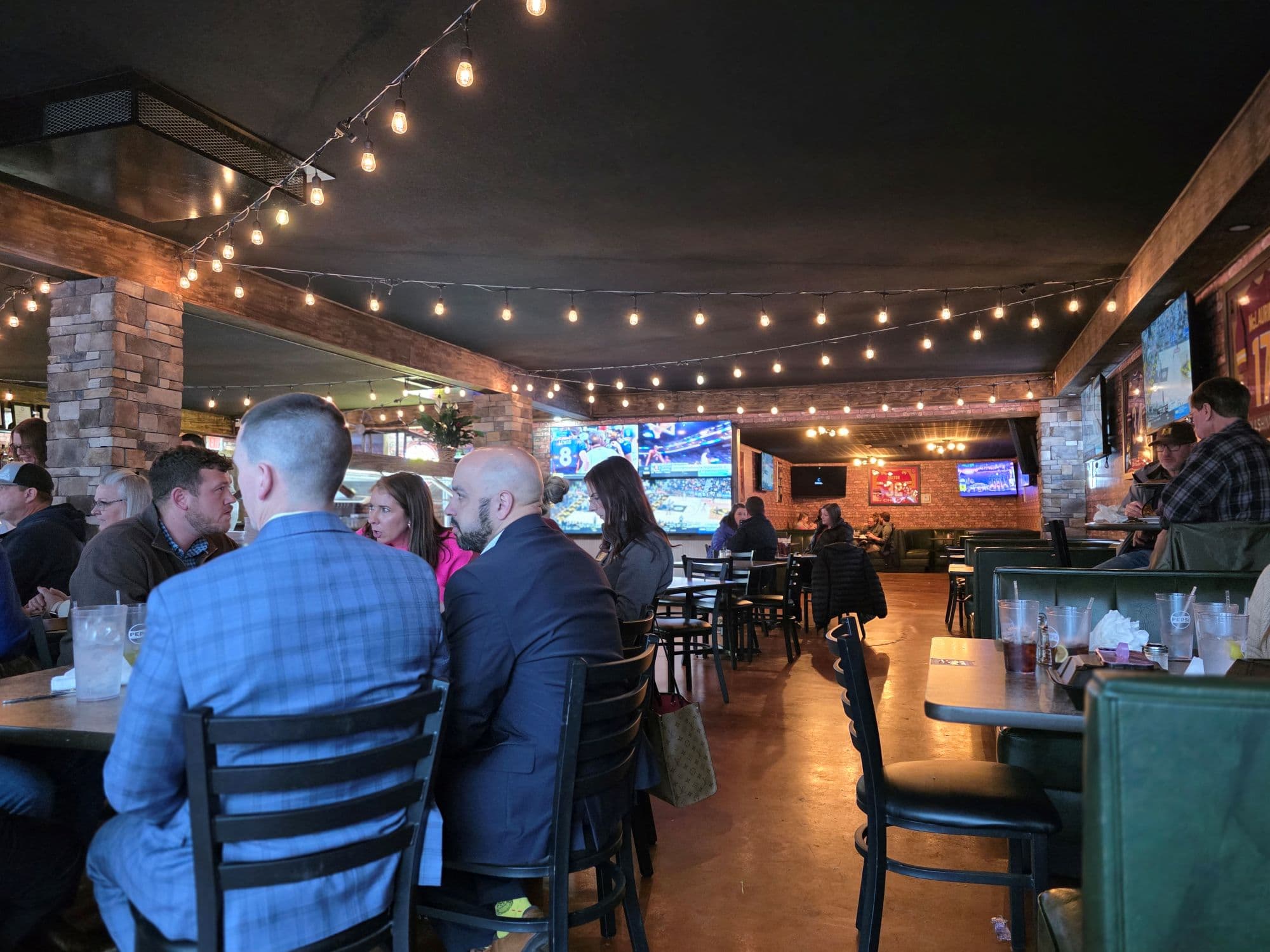 Interior of a lively restaurant with patrons seated at tables and screens displaying sports events, illuminated by string lights.