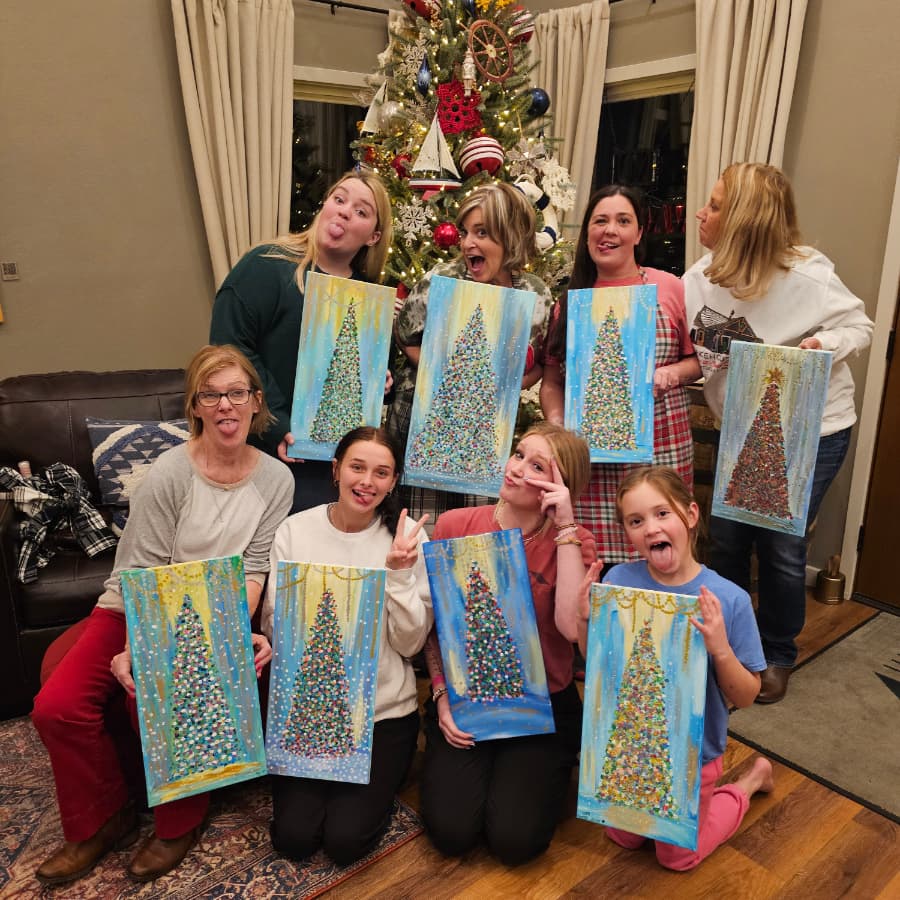 A group of eight women pose playfully in front of a decorated Christmas tree, holding their painted Christmas tree artwork.